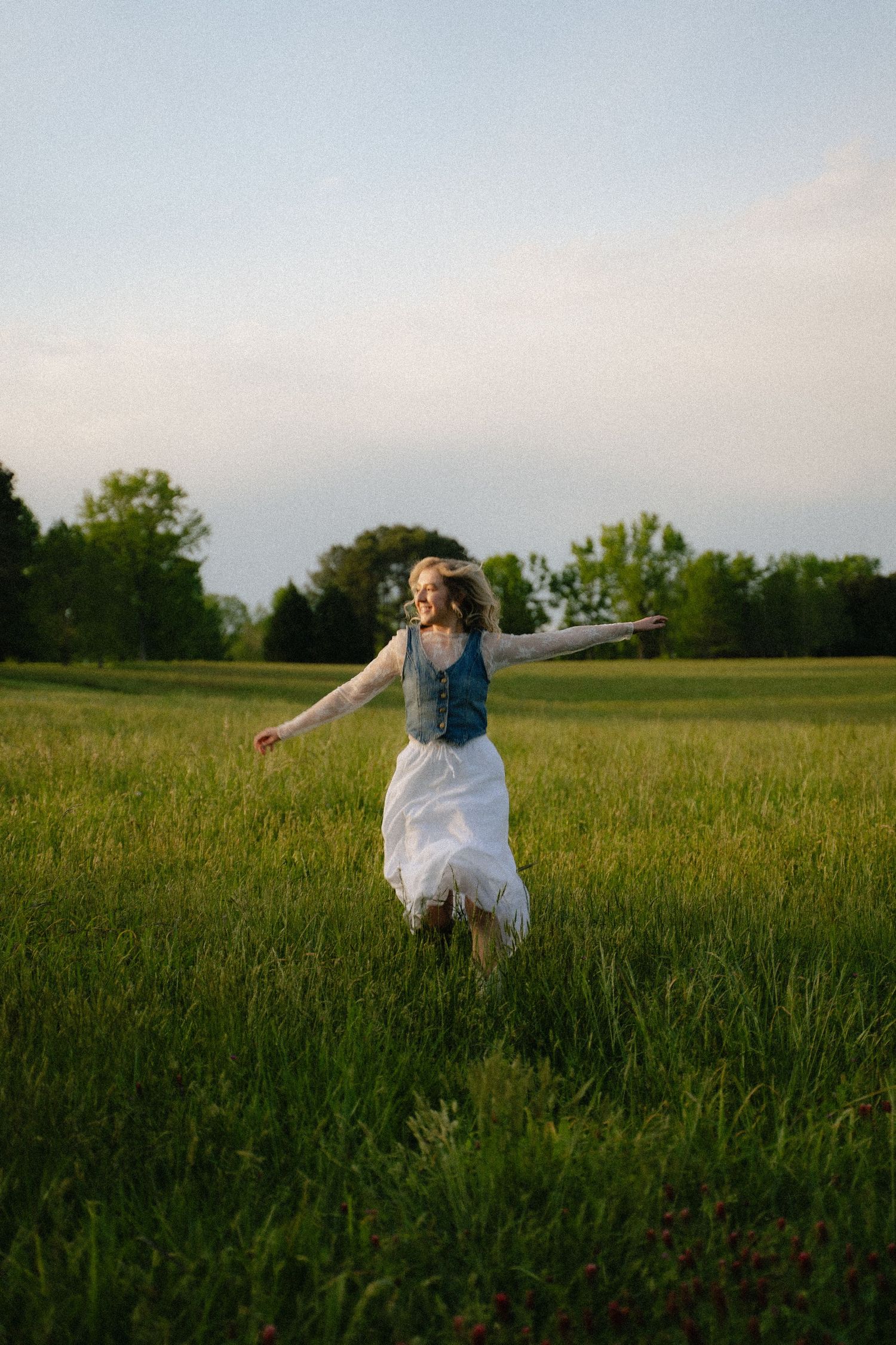 Joyful woman in white skirt and denim vest twirls in sunny green meadow at sunset.