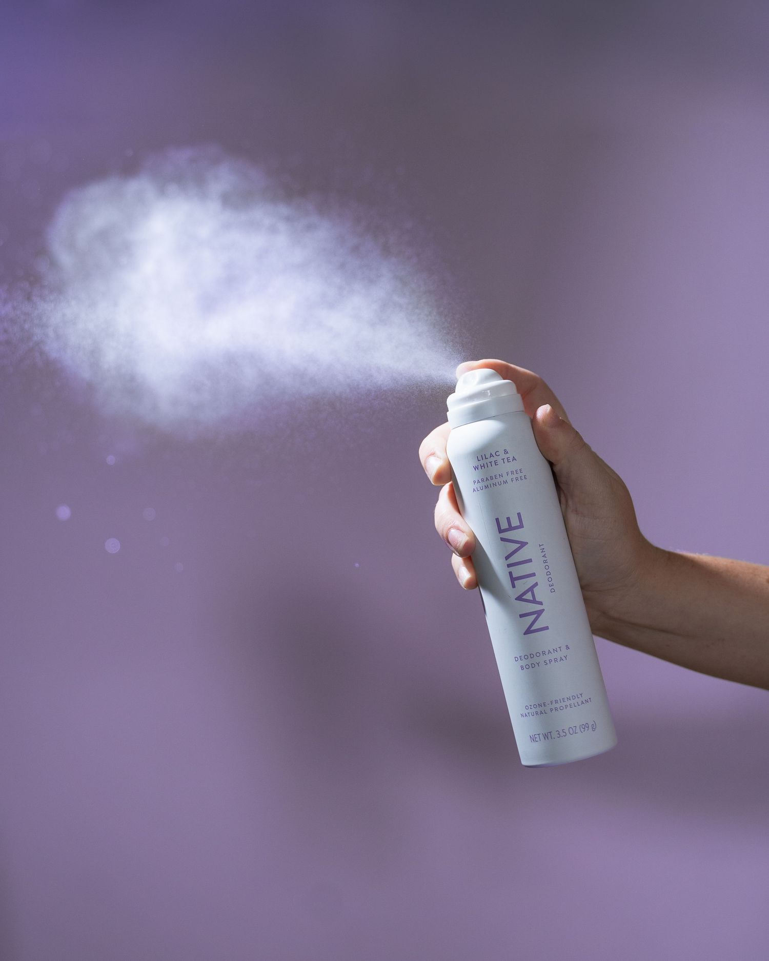 Hand spraying Native deodorant body spray with visible mist cloud against a purple background.
