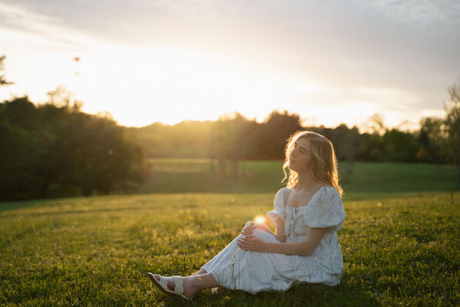 Young woman in white dress sitting on green grass during golden sunset in open field.
