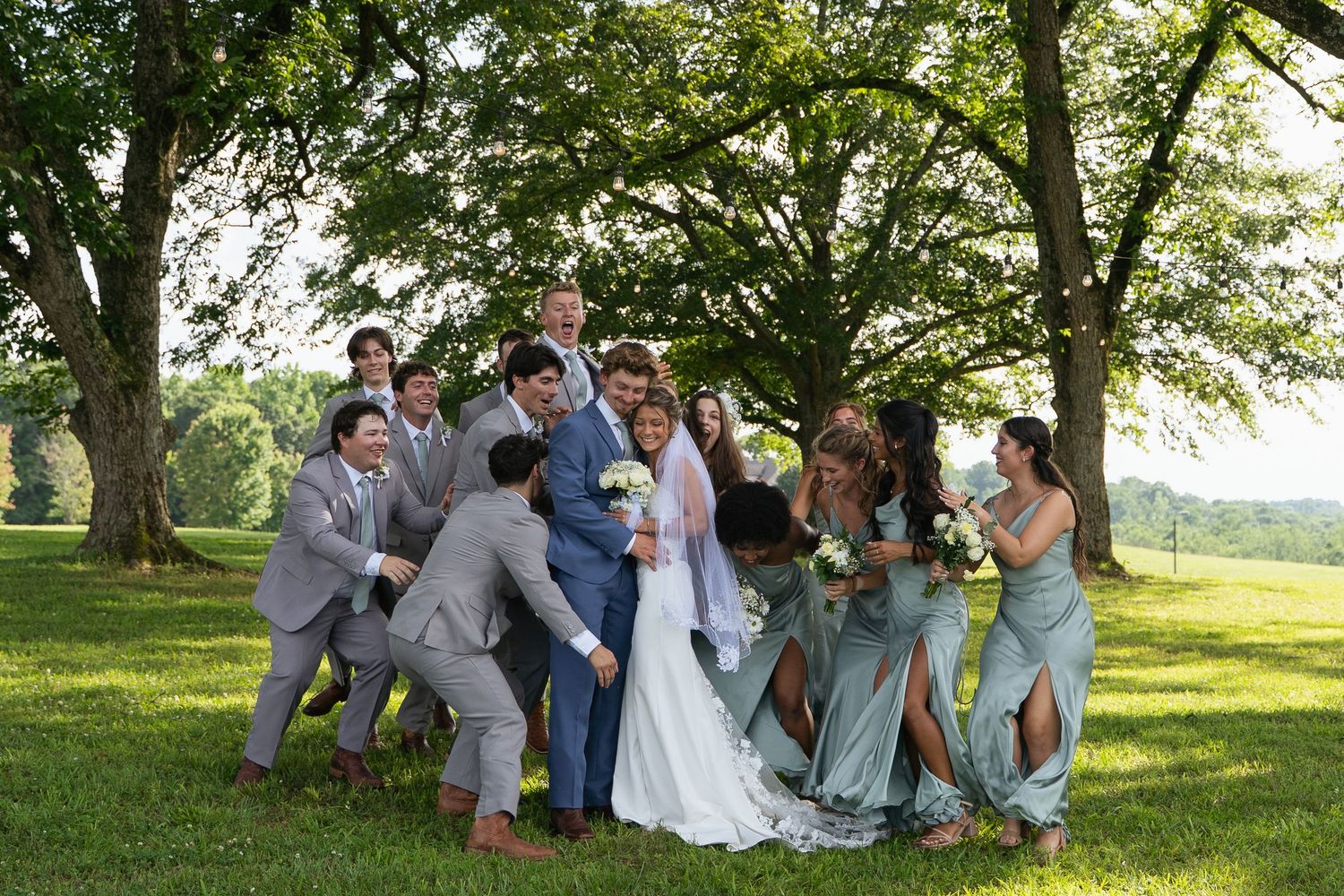 Bride and groom surrounded by cheerful wedding party in sage green and gray attire under large trees.