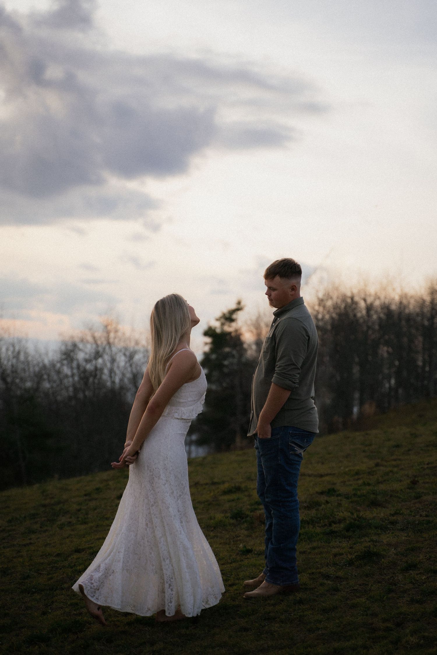 Couple holding hands at sunset on a grassy hill, woman in white lace dress, man in casual attire with cloudy sky backdrop.