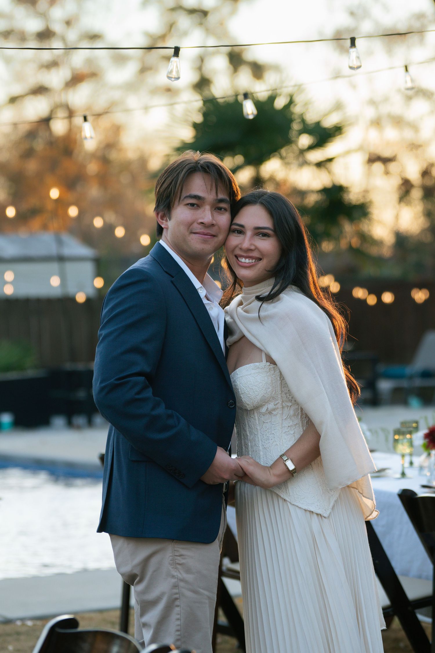 Smiling engaged couple embracing outdoors at golden hour with string lights behind them.