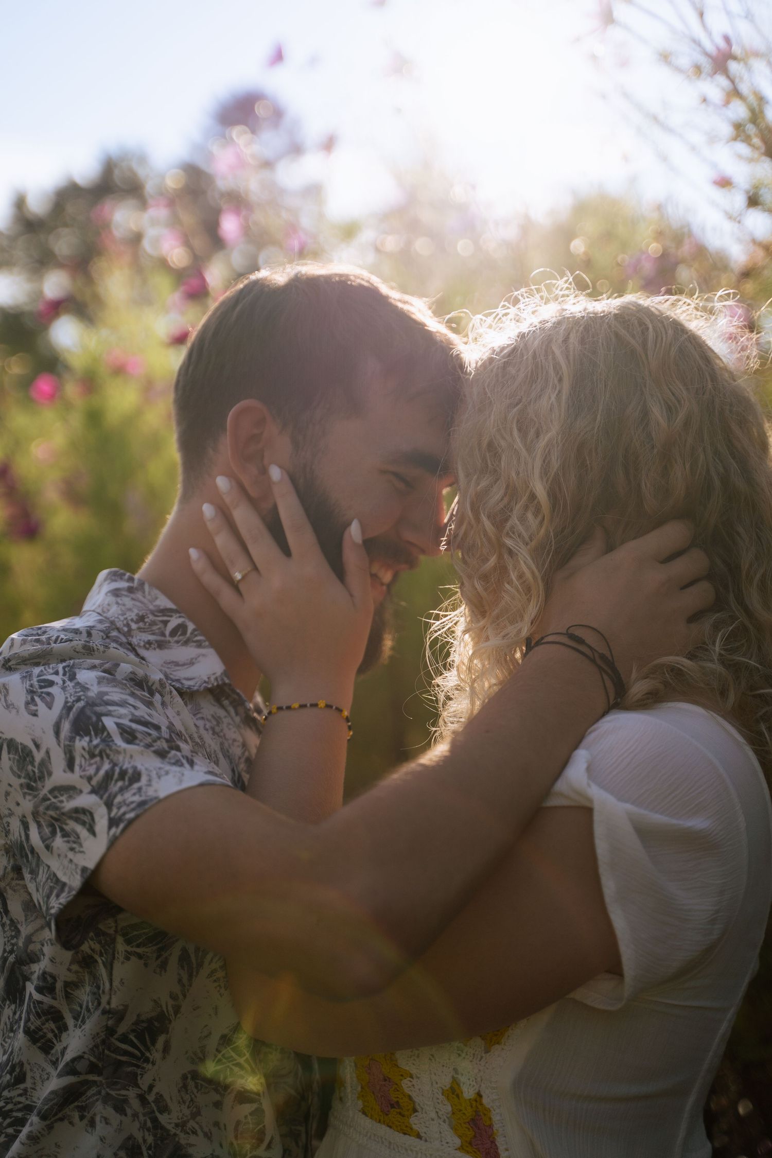 Couple sharing a romantic moment in sunlit flower field, foreheads touching, hands on each other's faces.
