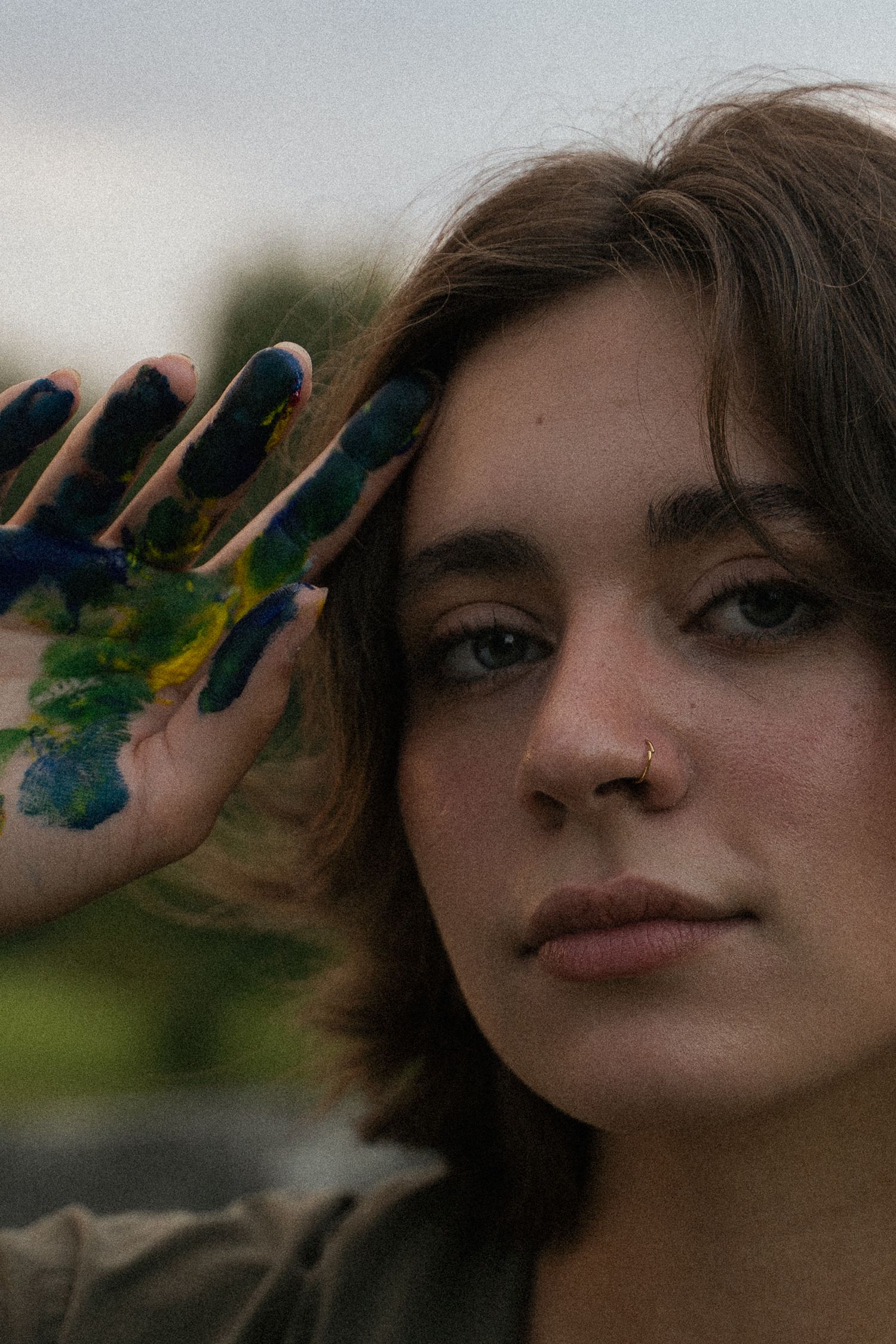 Close-up portrait of young woman with paint-covered hand raised beside her face outdoors.
