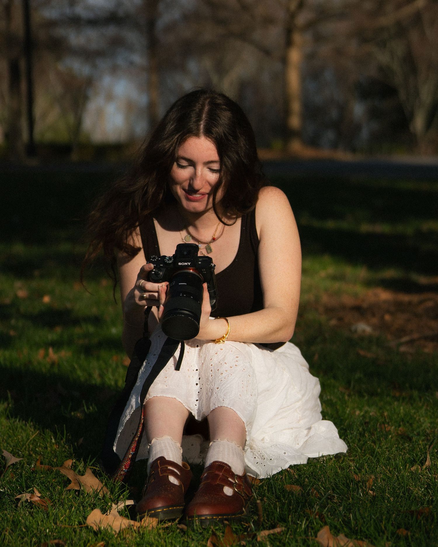 Smiling young woman sitting on grass in a park, reviewing photos on a Sony camera in golden sunlight.