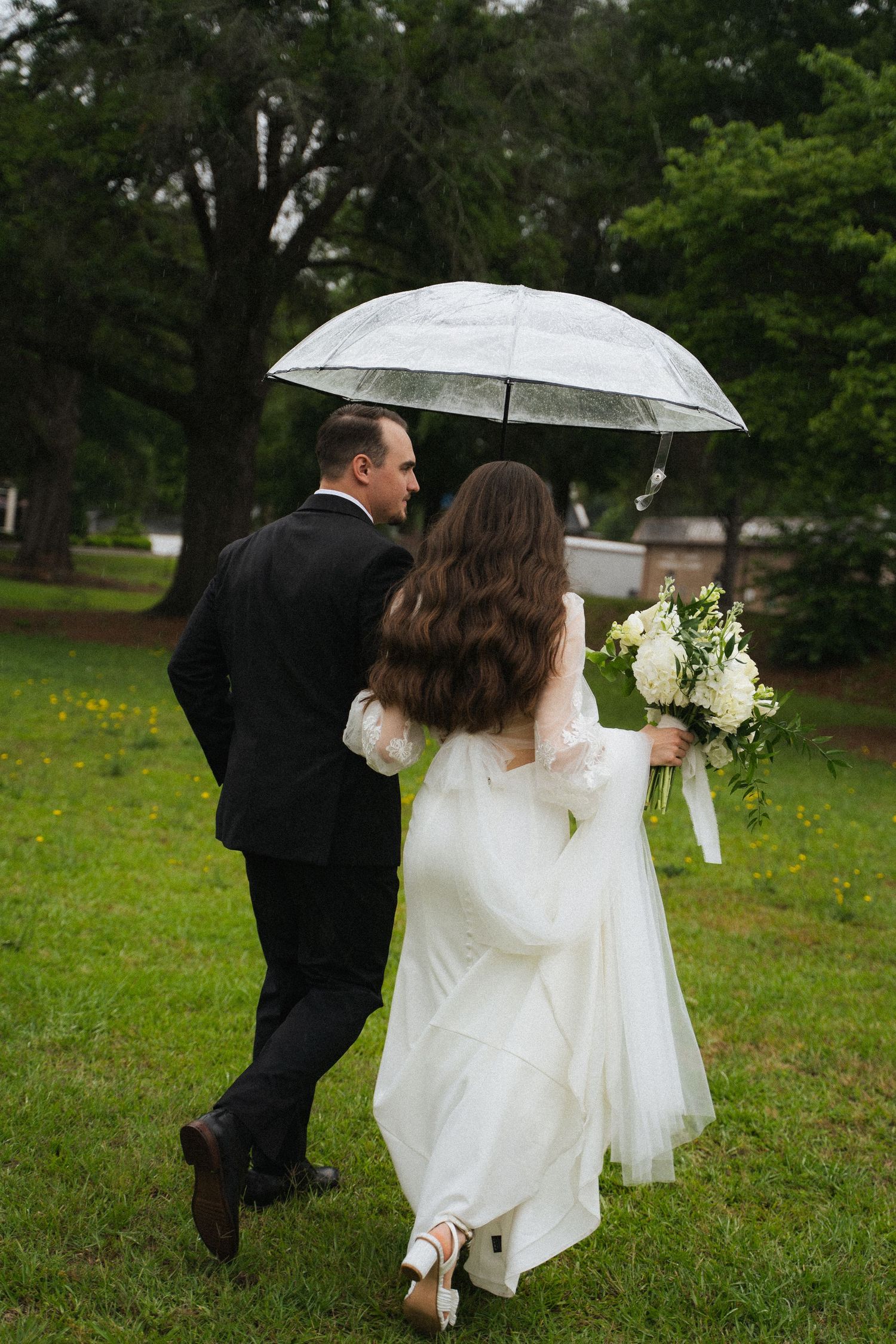 Bride and groom walking under clear umbrella in rain through lush green park.
