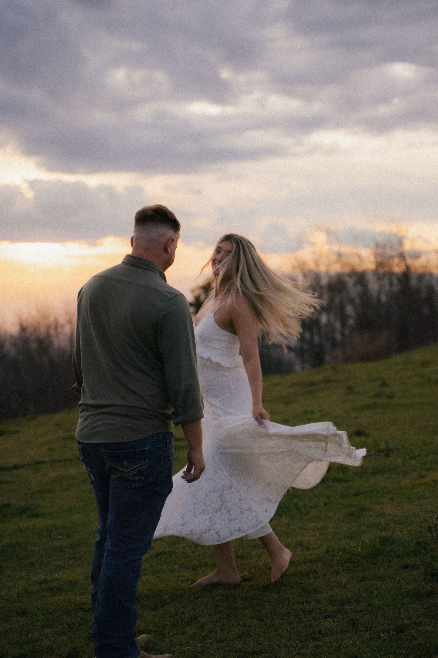 Couple dancing at sunset on grassy hilltop, woman in white dress with hair blowing in wind.