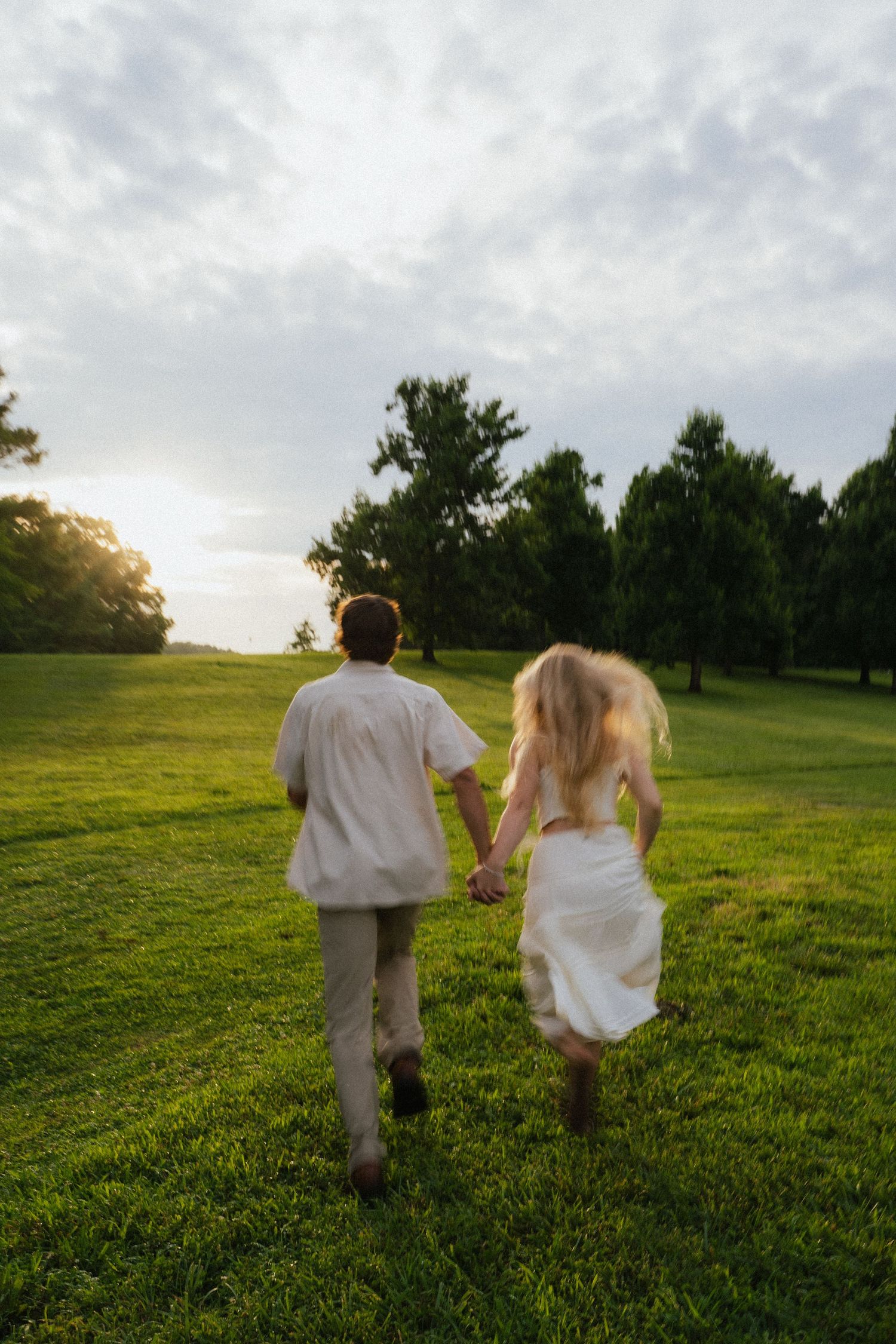 Couple runs hand in hand across a lush green park lawn toward a glowing sunset.