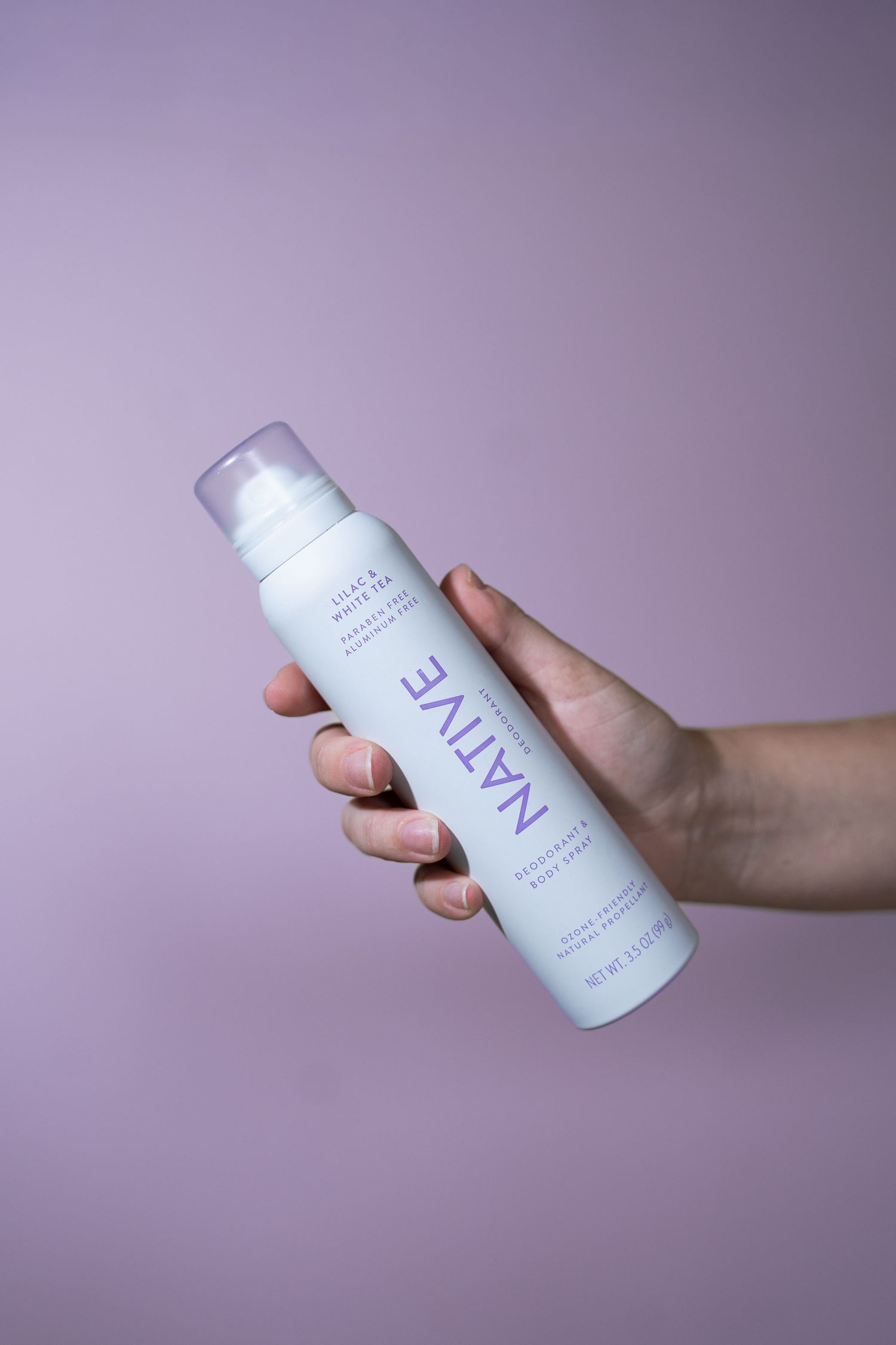 Hand holding Native lilac and white tea deodorant body spray against a soft purple background.