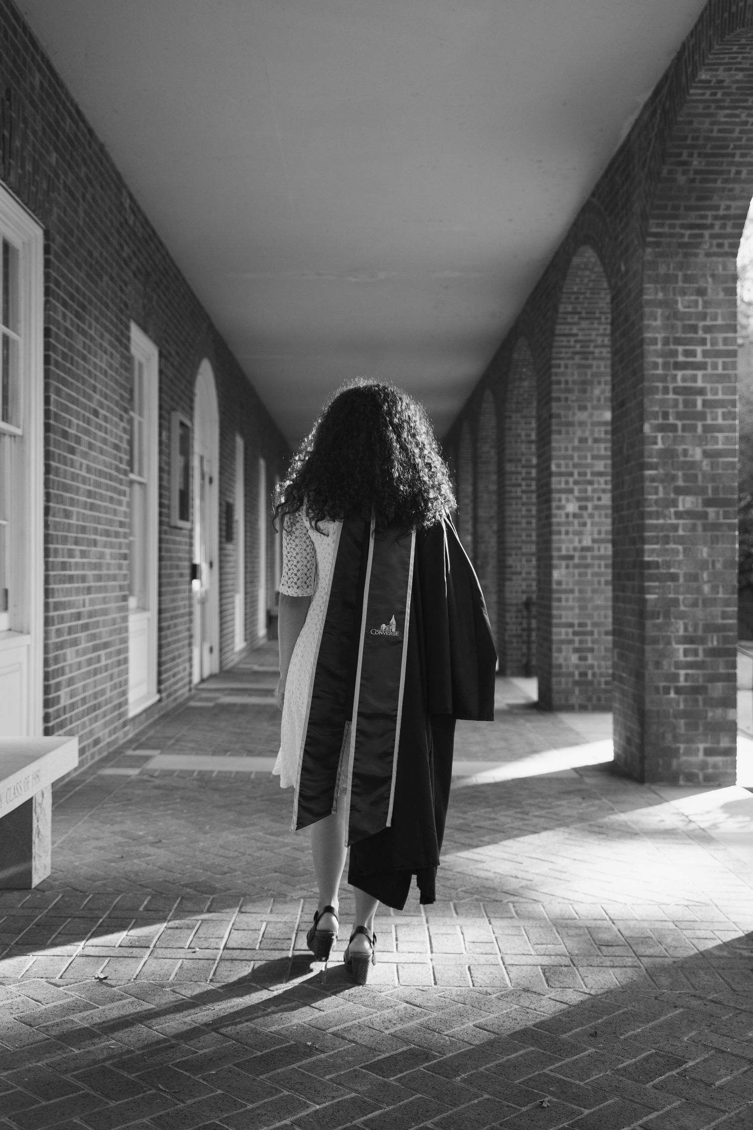 Black and white photo of graduate in cap and gown walking through brick archway corridor.
