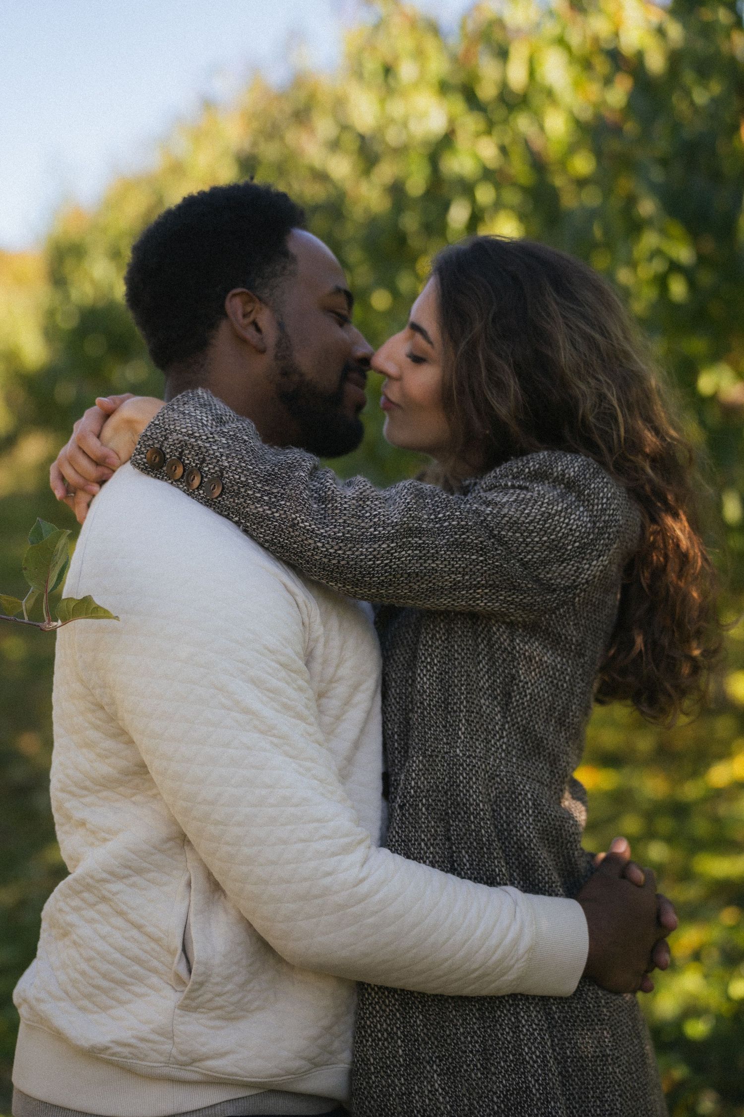 Interracial couple sharing a romantic embrace and almost kissing outdoors among lush green foliage.