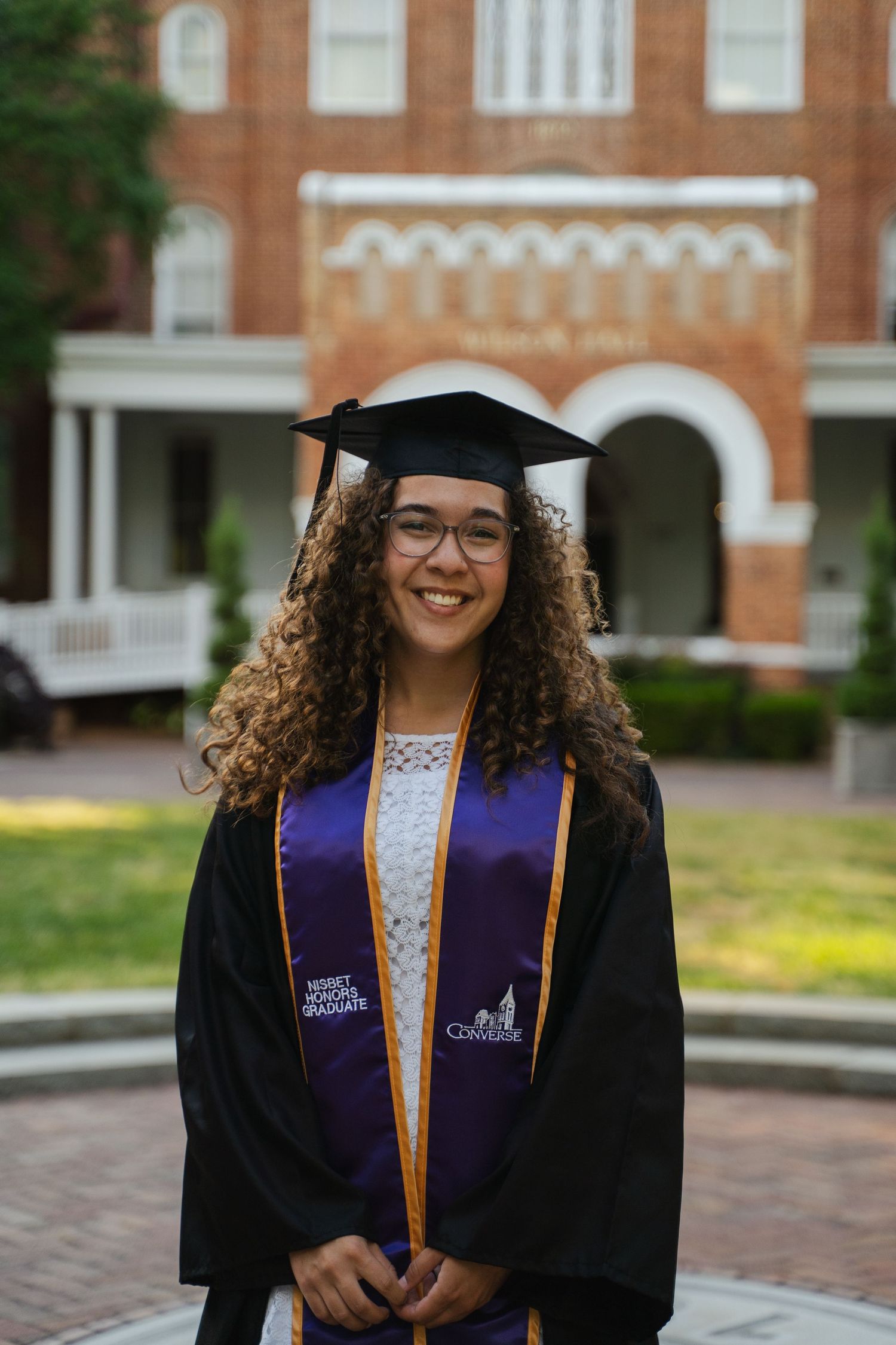 Smiling graduate in cap and gown with purple honor stole standing in front of a brick university building.