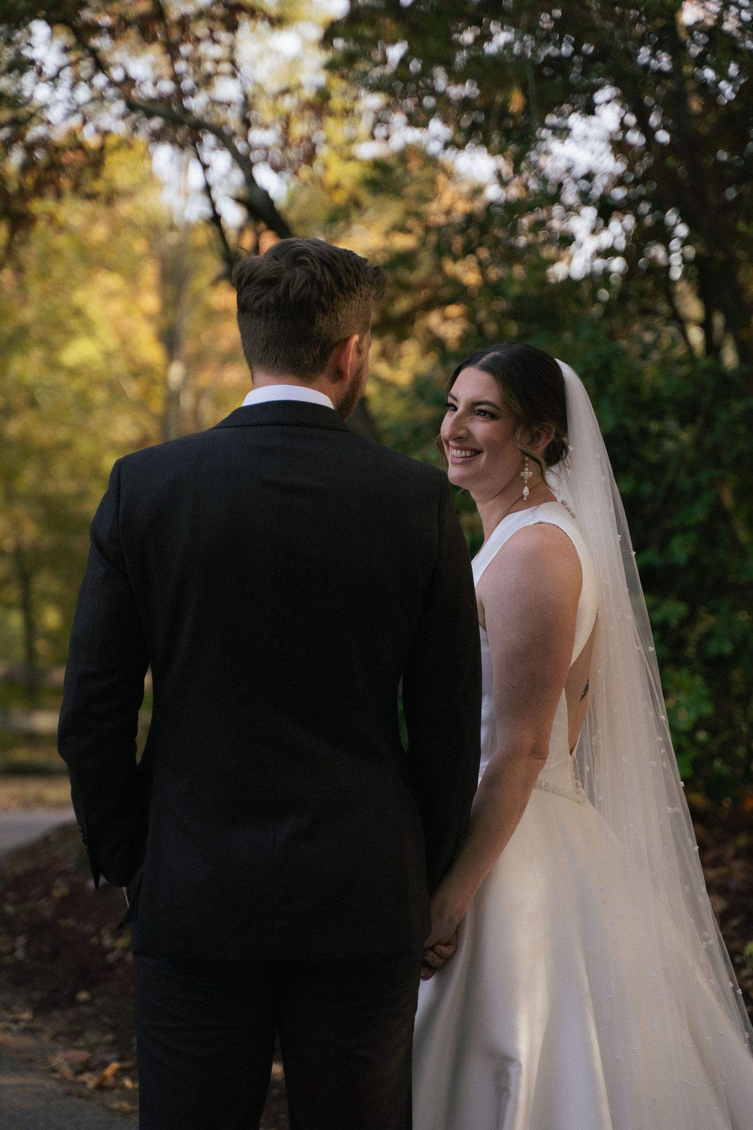 Smiling bride in white gown and veil walks with groom through tree-lined autumn path.