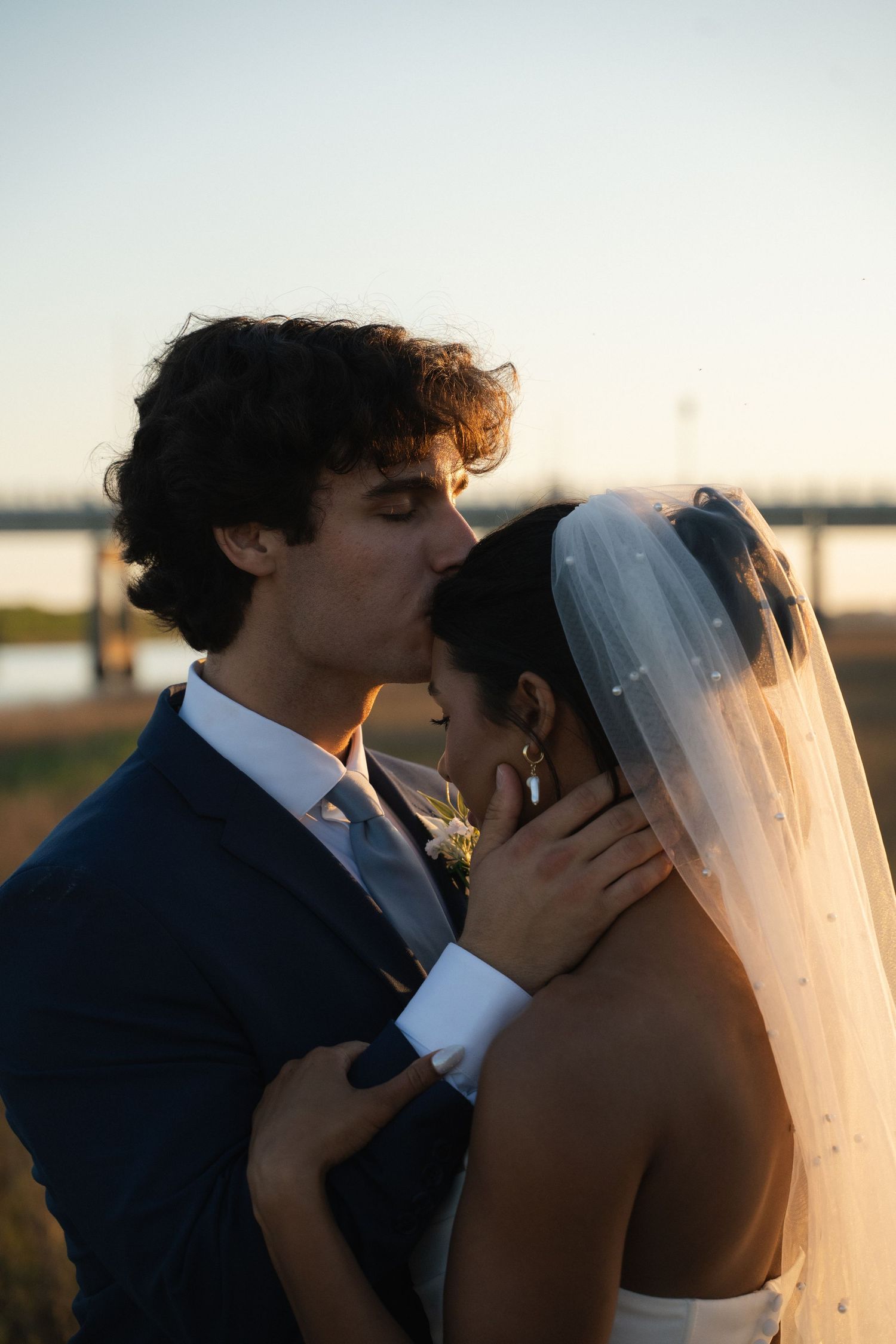 Groom in navy suit kisses bride in white veil tenderly at golden hour near waterfront.