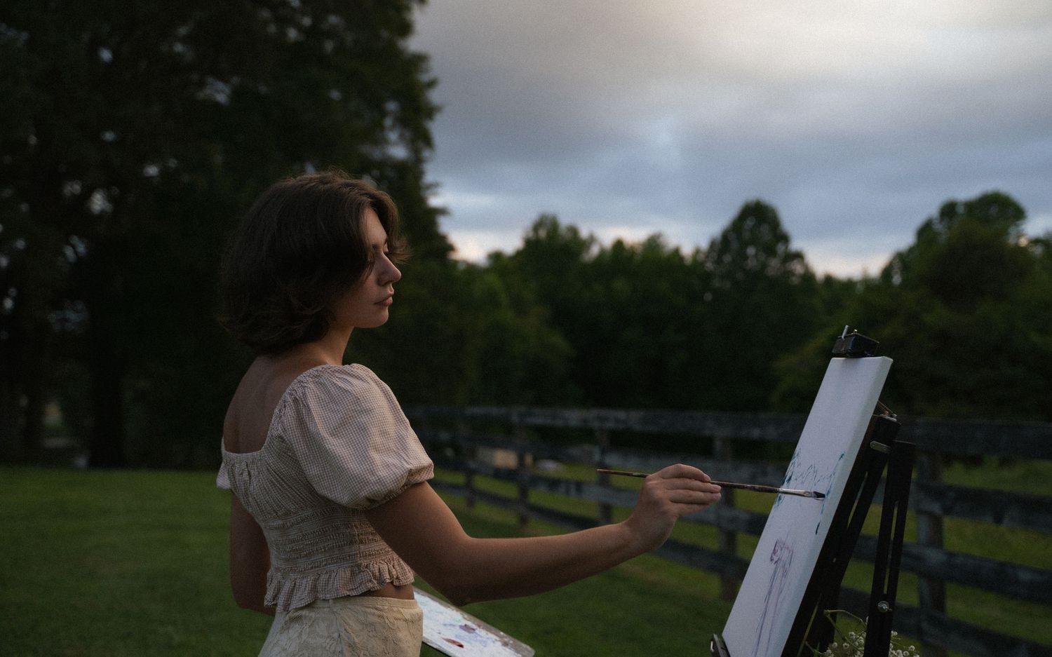 Young woman painting on canvas easel outdoors at dusk in a pastoral farm setting.