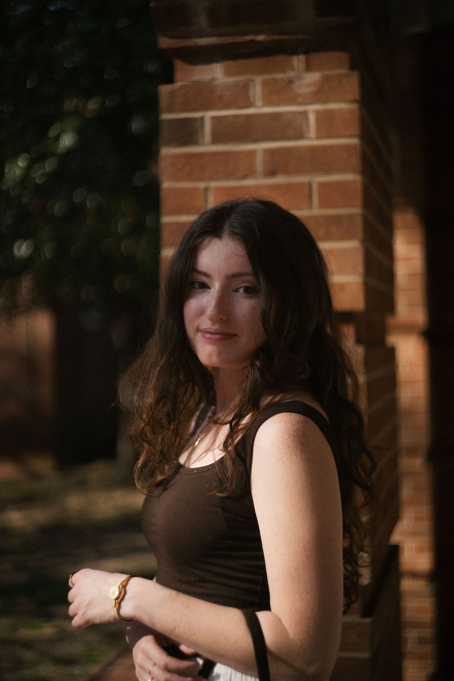 Young brunette woman smiling softly near a brick pillar in warm sunlight, wearing a dark sleeveless top.