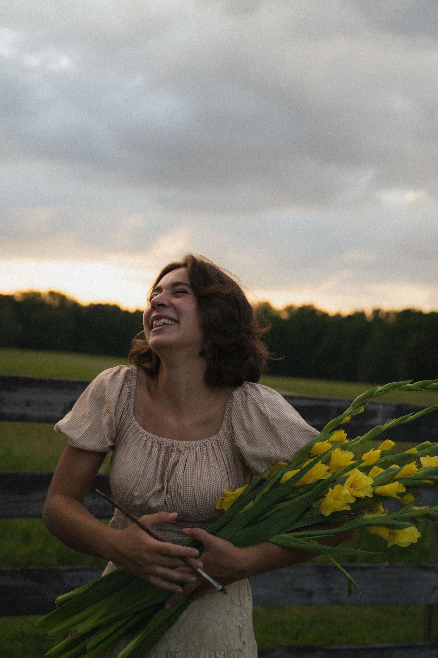 a young woman cradles flowers in her arms and laughs in a grassy field at sunset