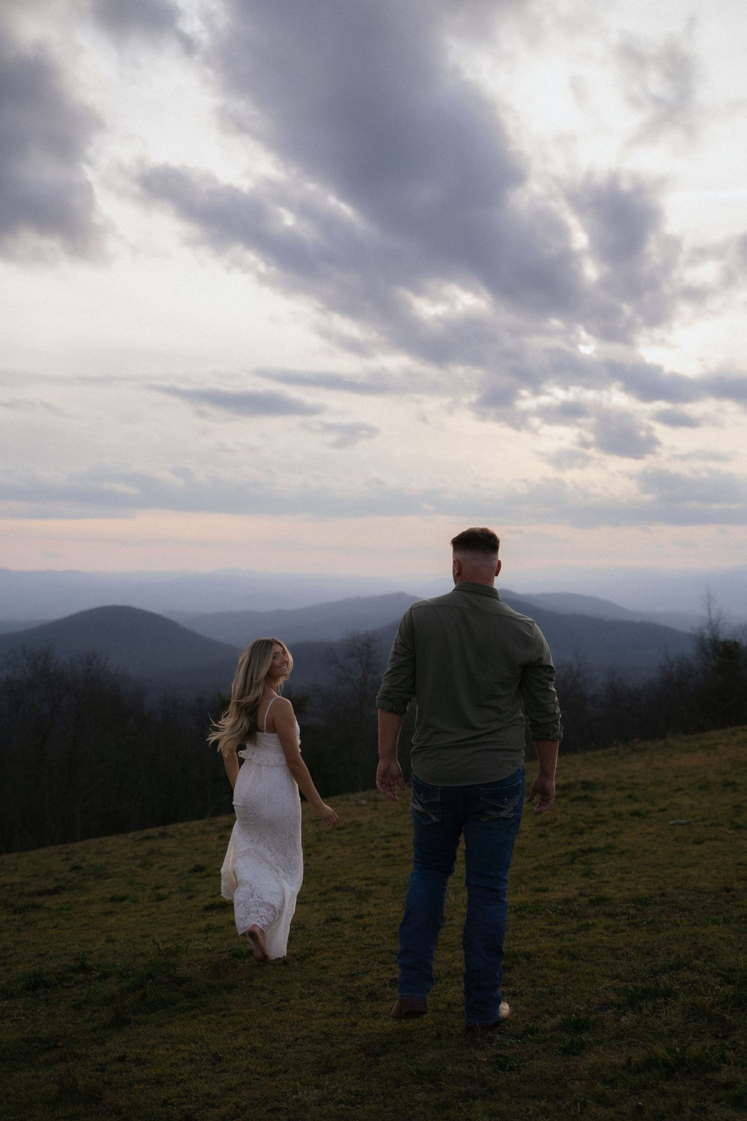 Couple walking on grassy hilltop at dusk, woman in white dress, man in casual wear, mountain vista behind.
