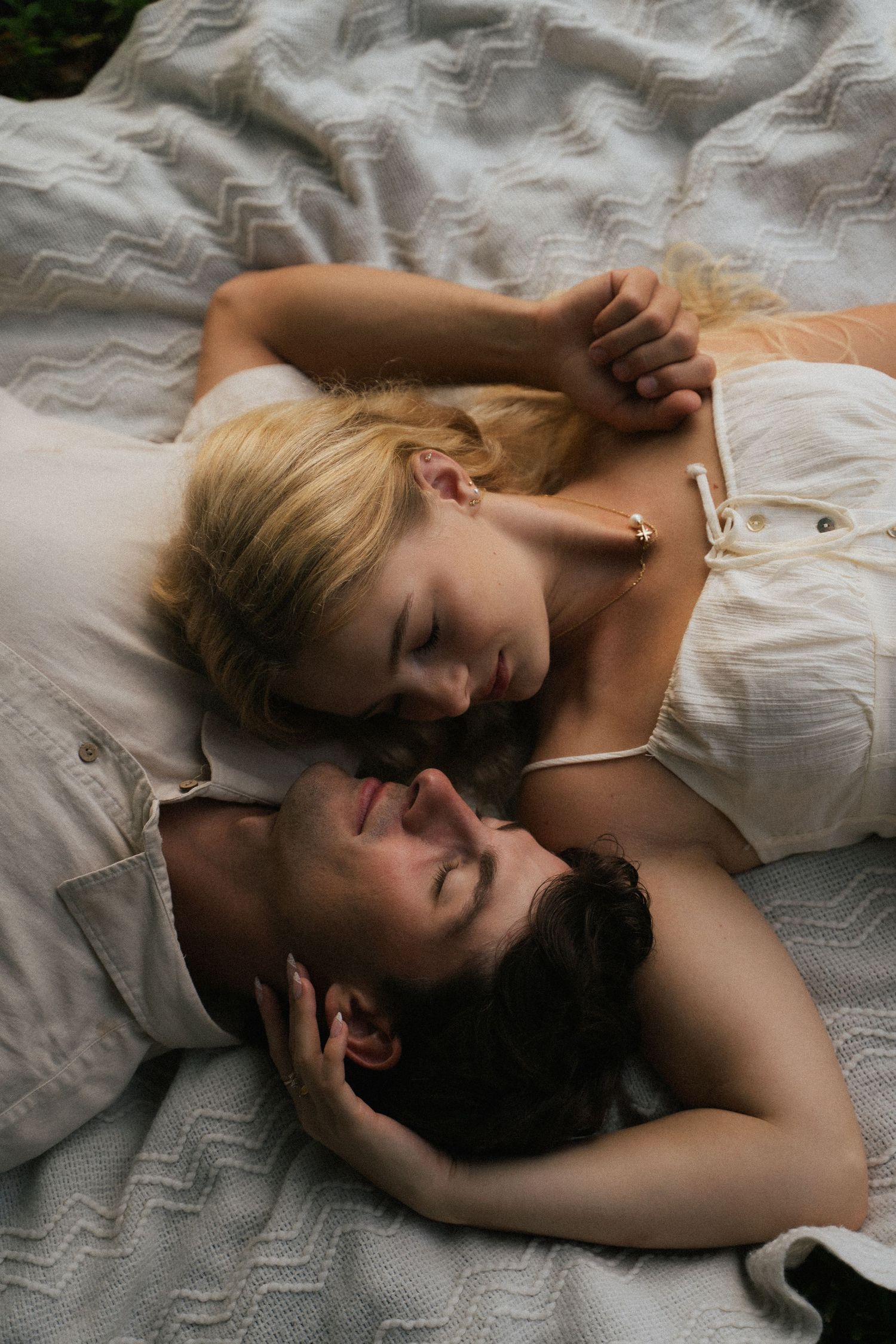 Couple lies together on a white blanket outdoors, sharing a tender intimate moment.
