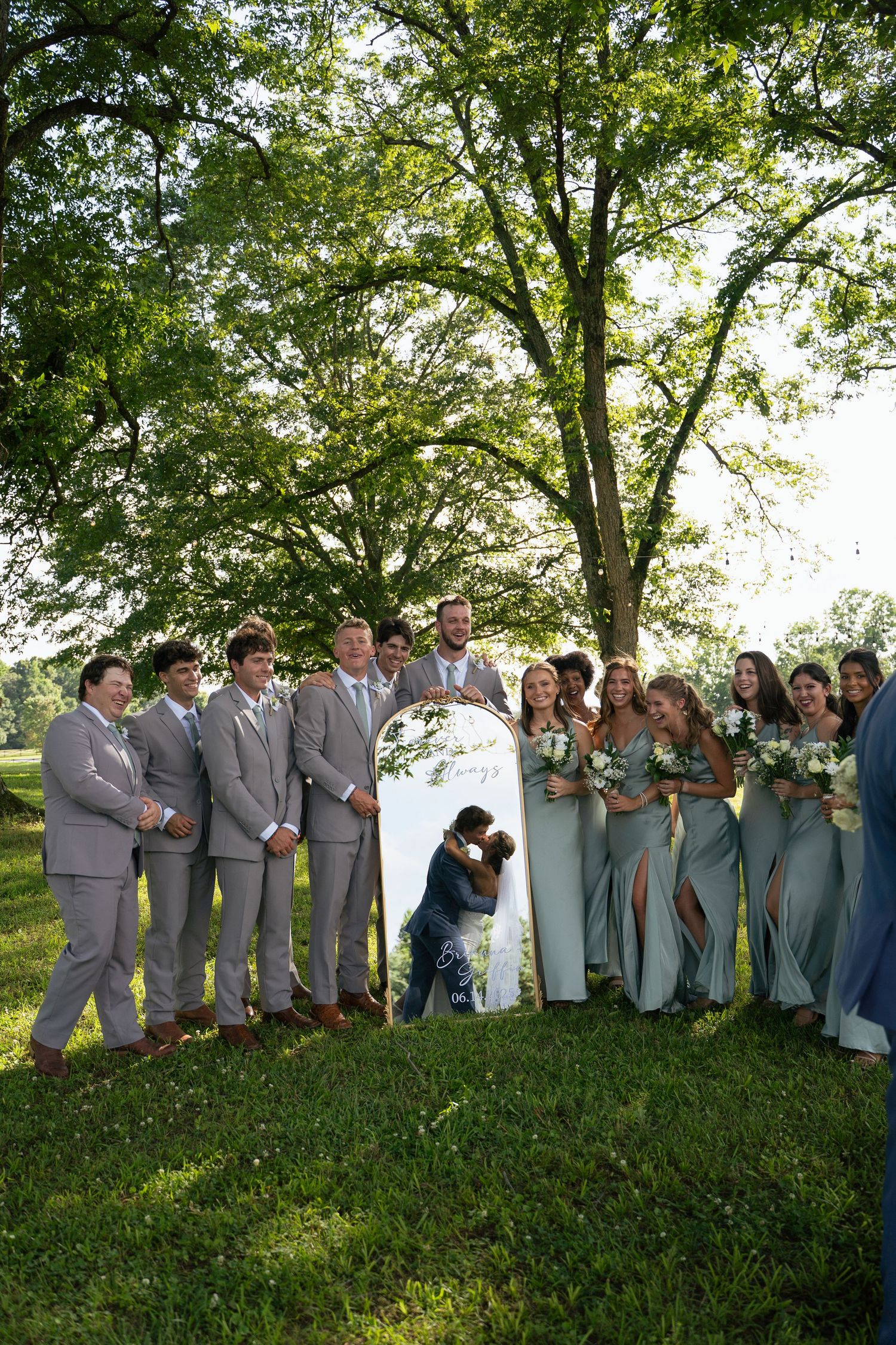 Wedding party poses outdoors under large tree with kissing couple behind decorative sign.