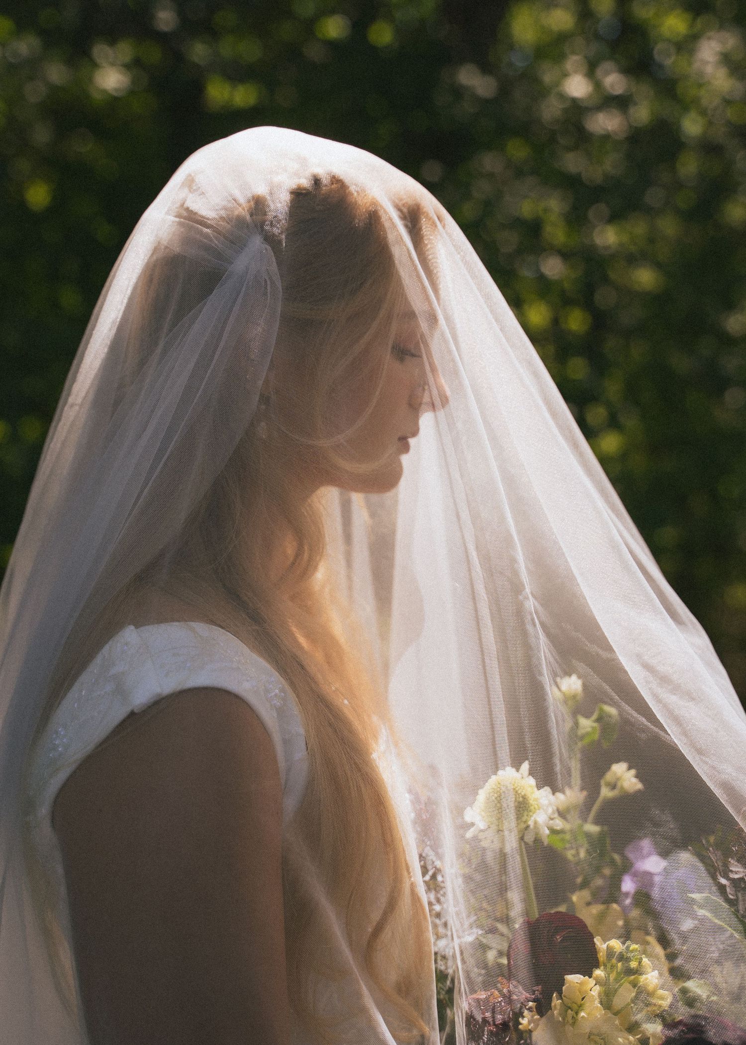 Ethereal bride in white veil holding floral bouquet in soft natural light.