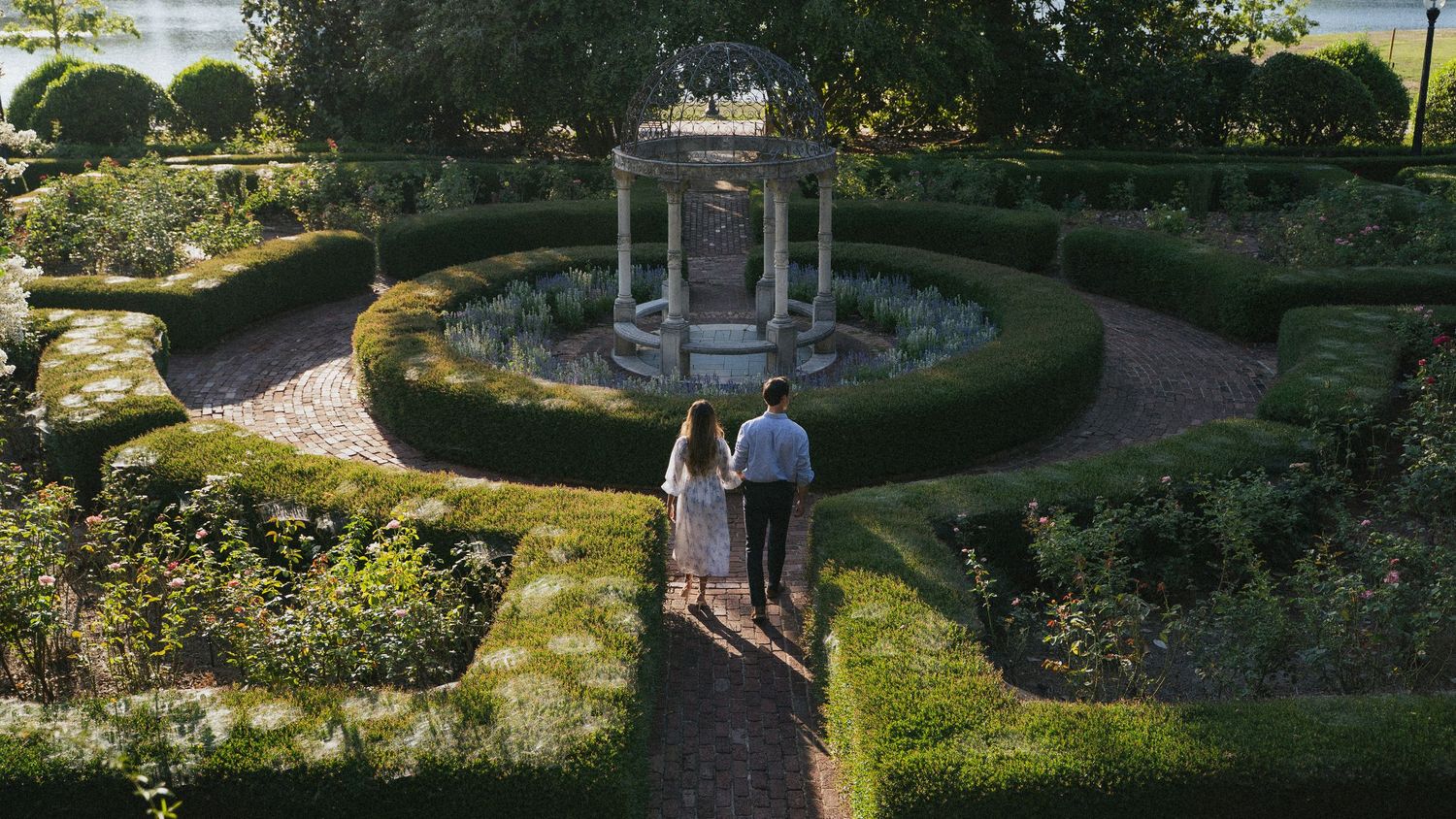 Couple walking hand in hand through a formal garden with manicured hedges and a stone gazebo at sunset.