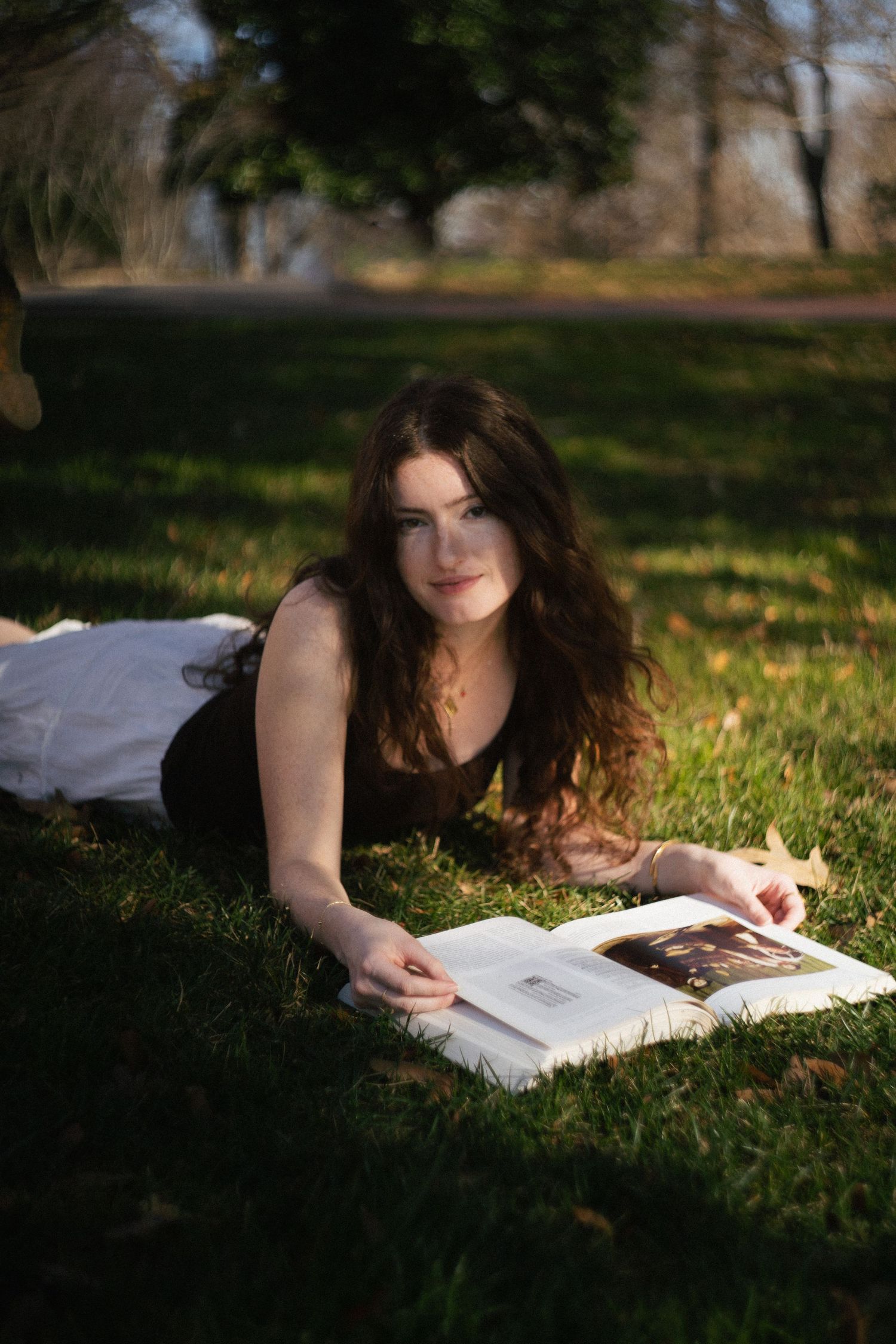 Beautiful brunette woman lying on green grass in a sunny park, looking up while reading an open book.