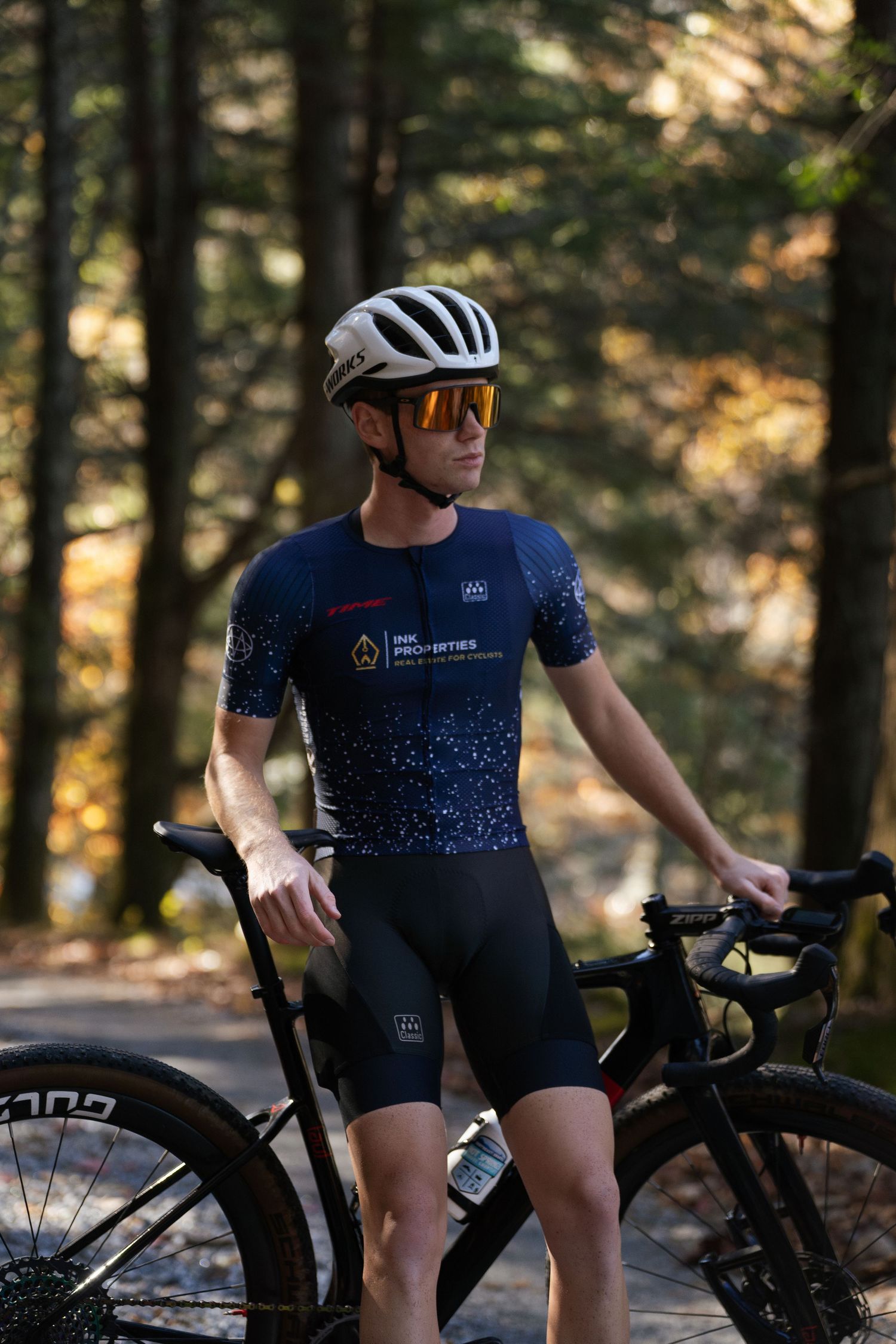 Male cyclist in navy racing kit and helmet standing with road bike in autumn forest.