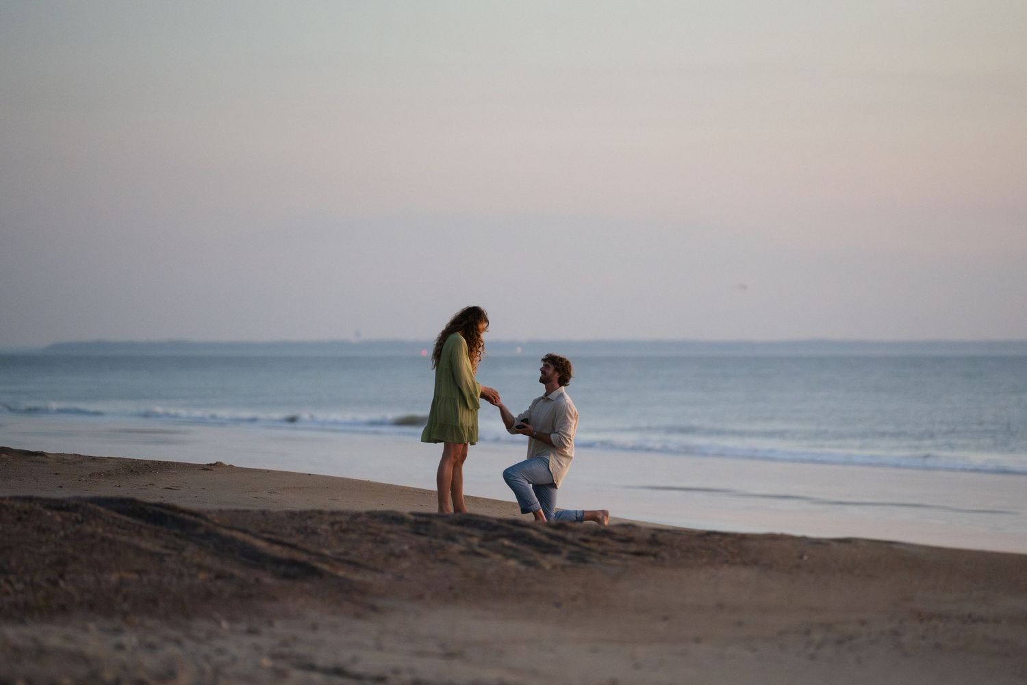 Man proposing to woman on a beach at sunset, kneeling in the sand with ocean waves in the background.