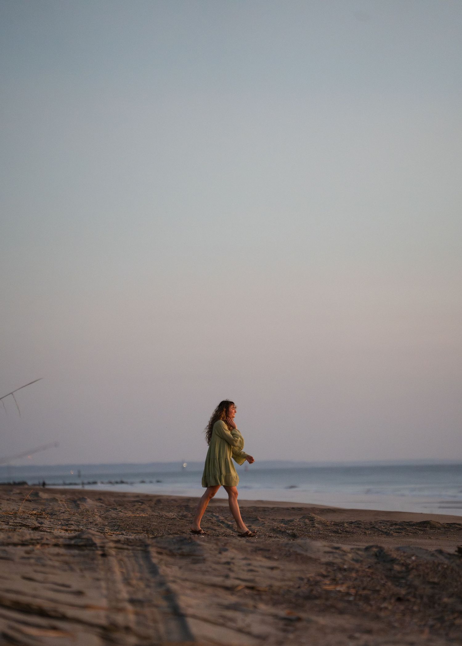 Woman in a green mini dress walking barefoot along a sandy beach at sunset with calm ocean behind her.