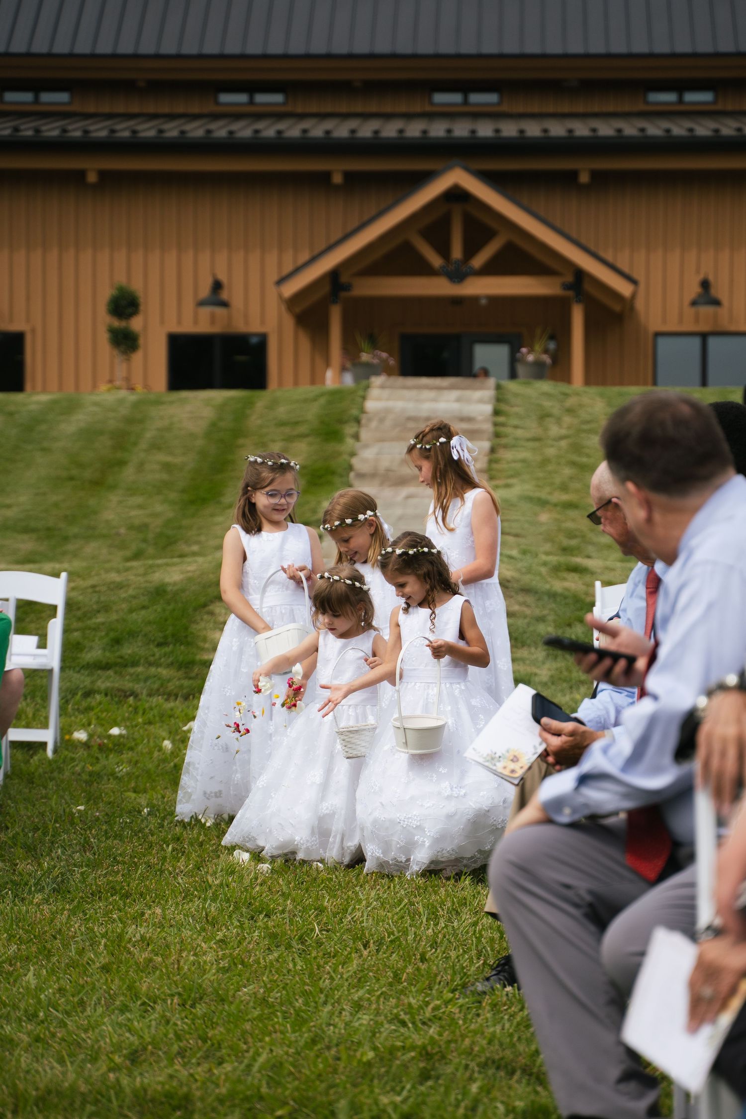 Five flower girls in white dresses and floral crowns scattering petals down the aisle at an outdoor wedding.