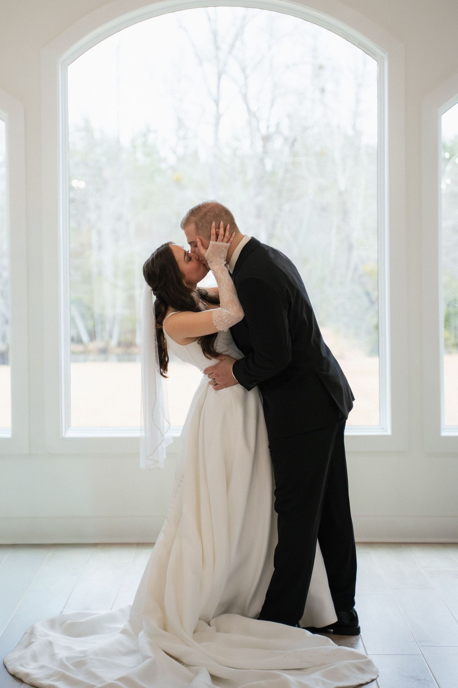 Bride and groom share first kiss in elegant white chapel with arched windows and natural light.