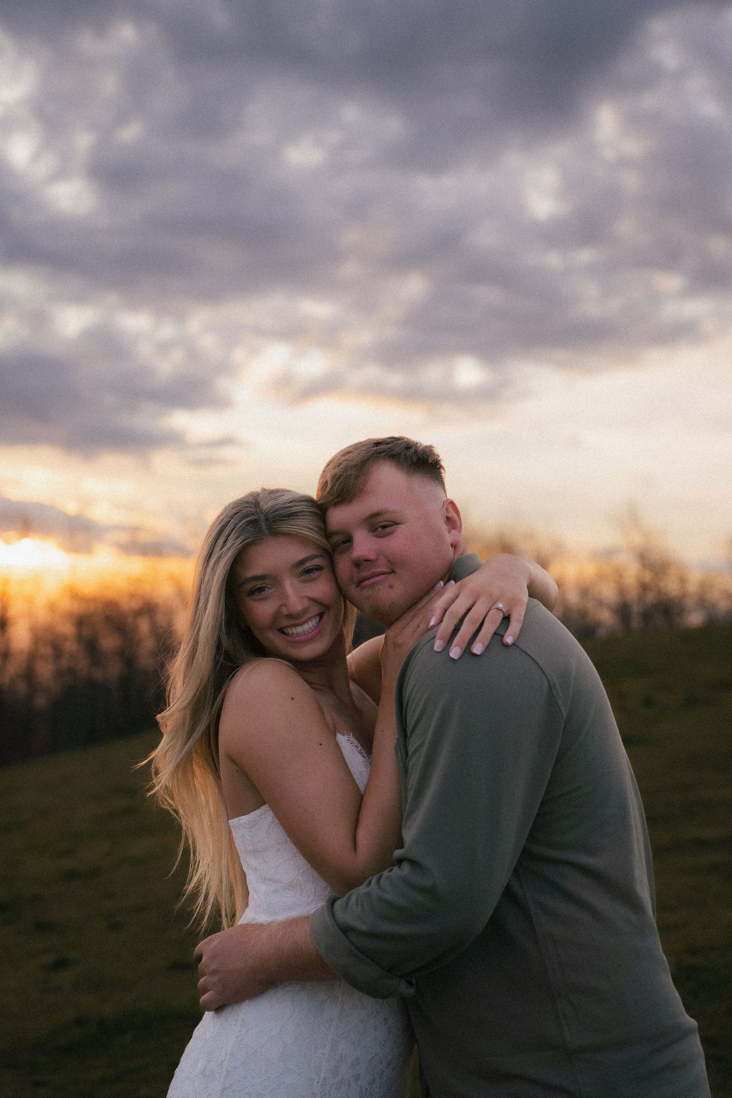 Smiling couple embracing at sunset on hillside, woman in white dress with arms around man.