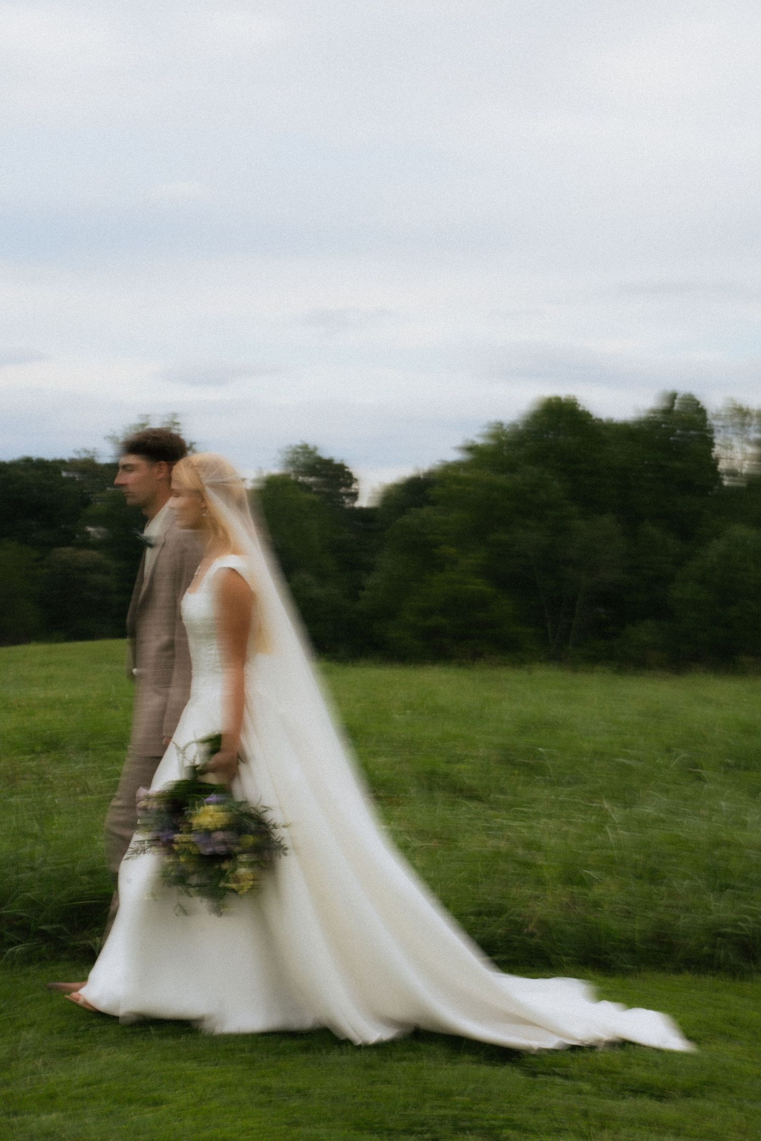 Bride and groom walking through green meadow, motion blur capturing ethereal wedding day moment.
