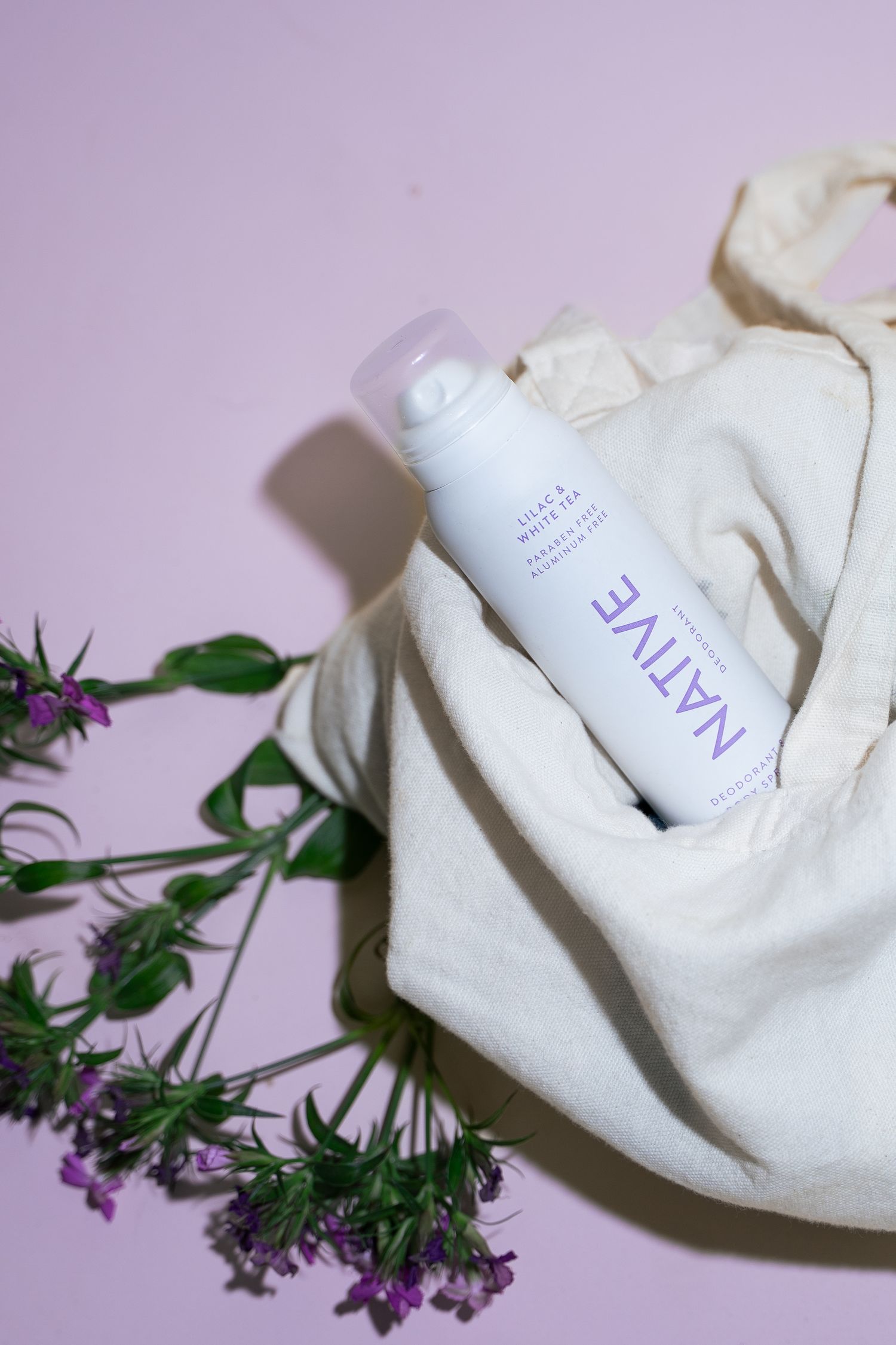 Native lilac and white tea deodorant body spray in a canvas tote bag with purple wildflowers on pink background.