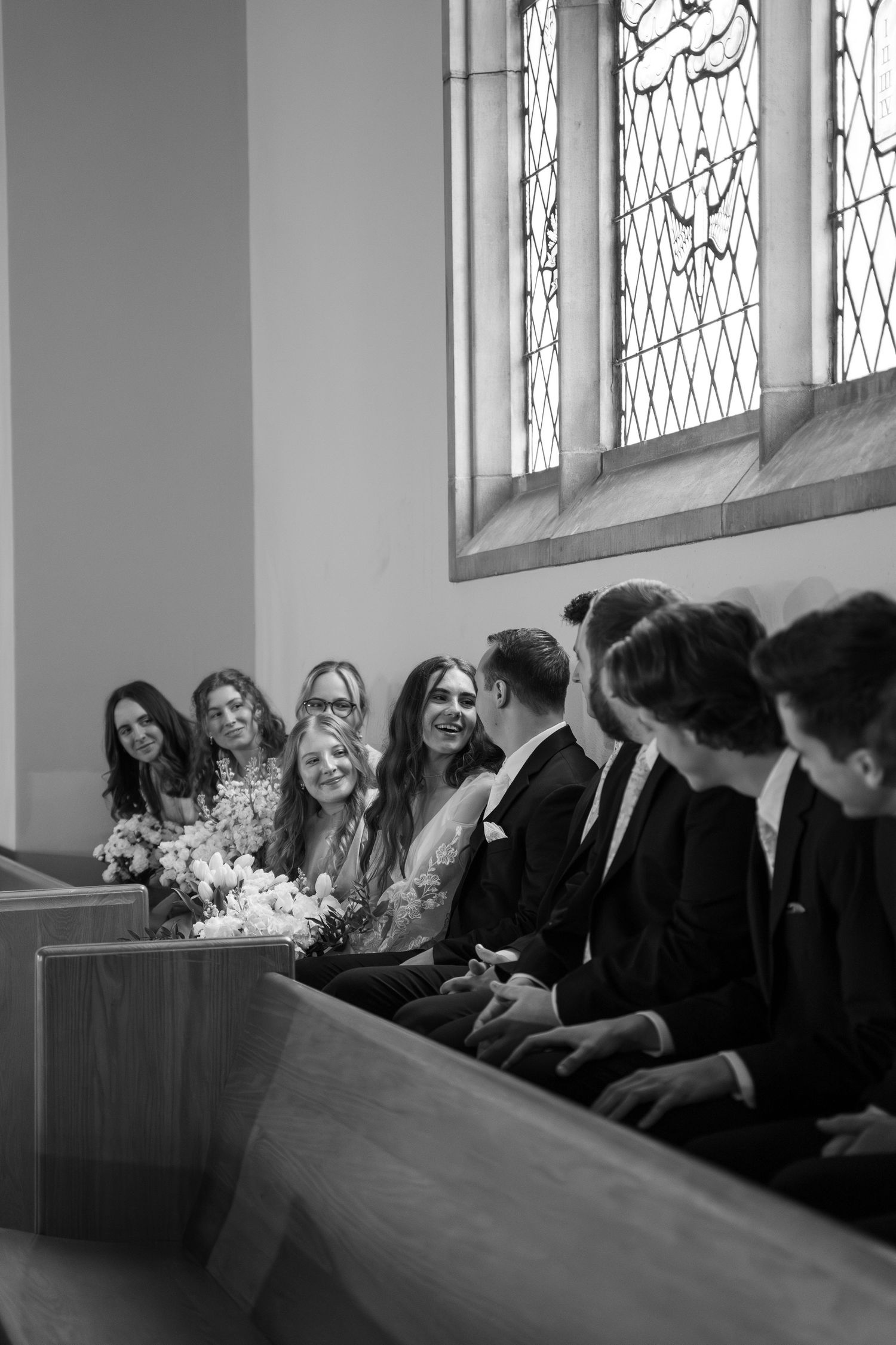 Black and white photo of wedding guests laughing together in church pews near stained glass windows.