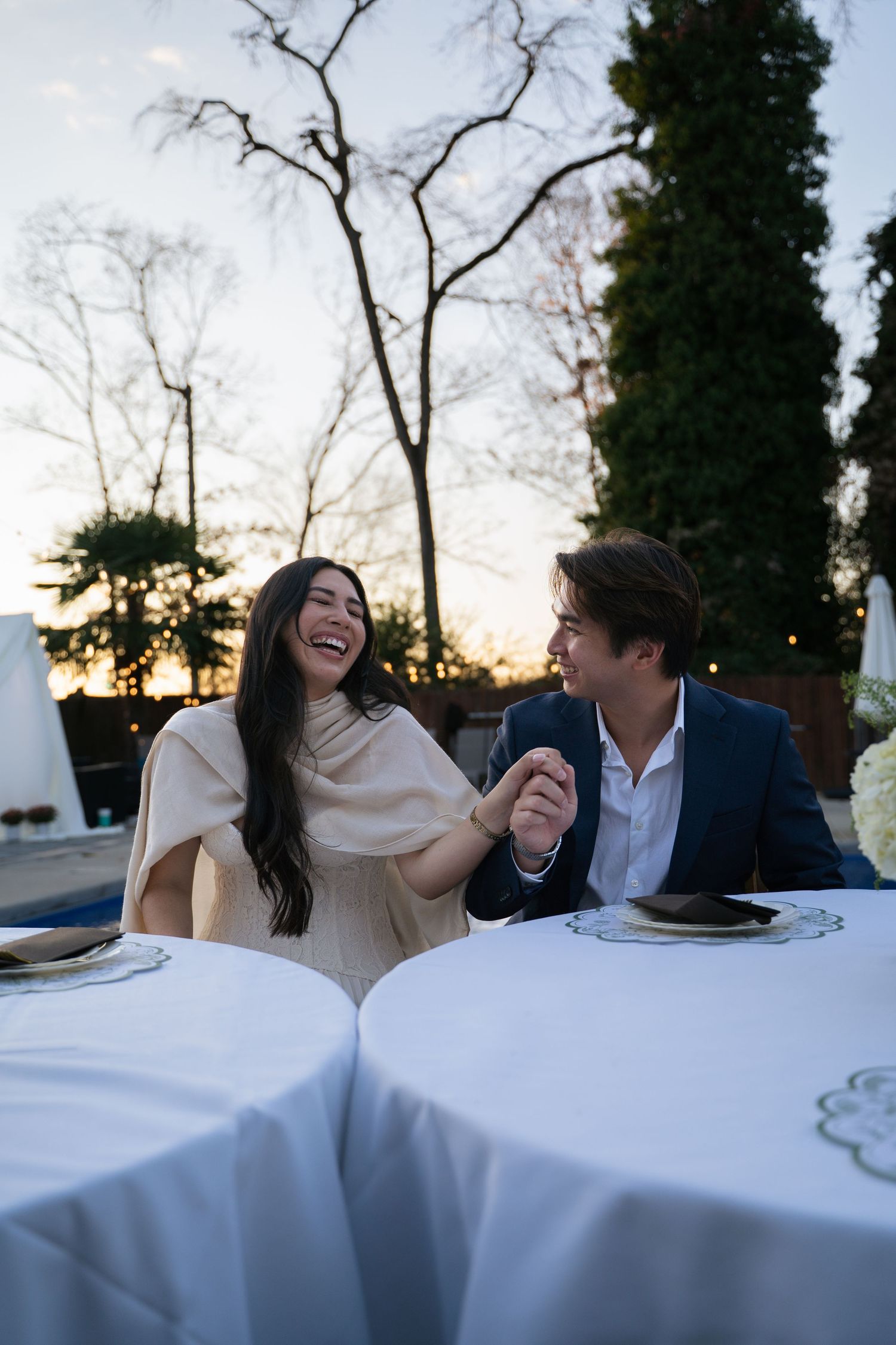 Couple holding hands and laughing at outdoor candlelit dinner table at sunset.
