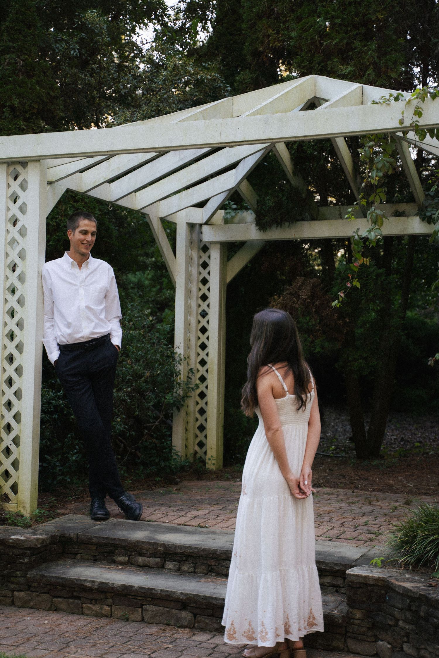 Couple poses under a white wooden pergola in a lush garden, man in white shirt smiling at woman.
