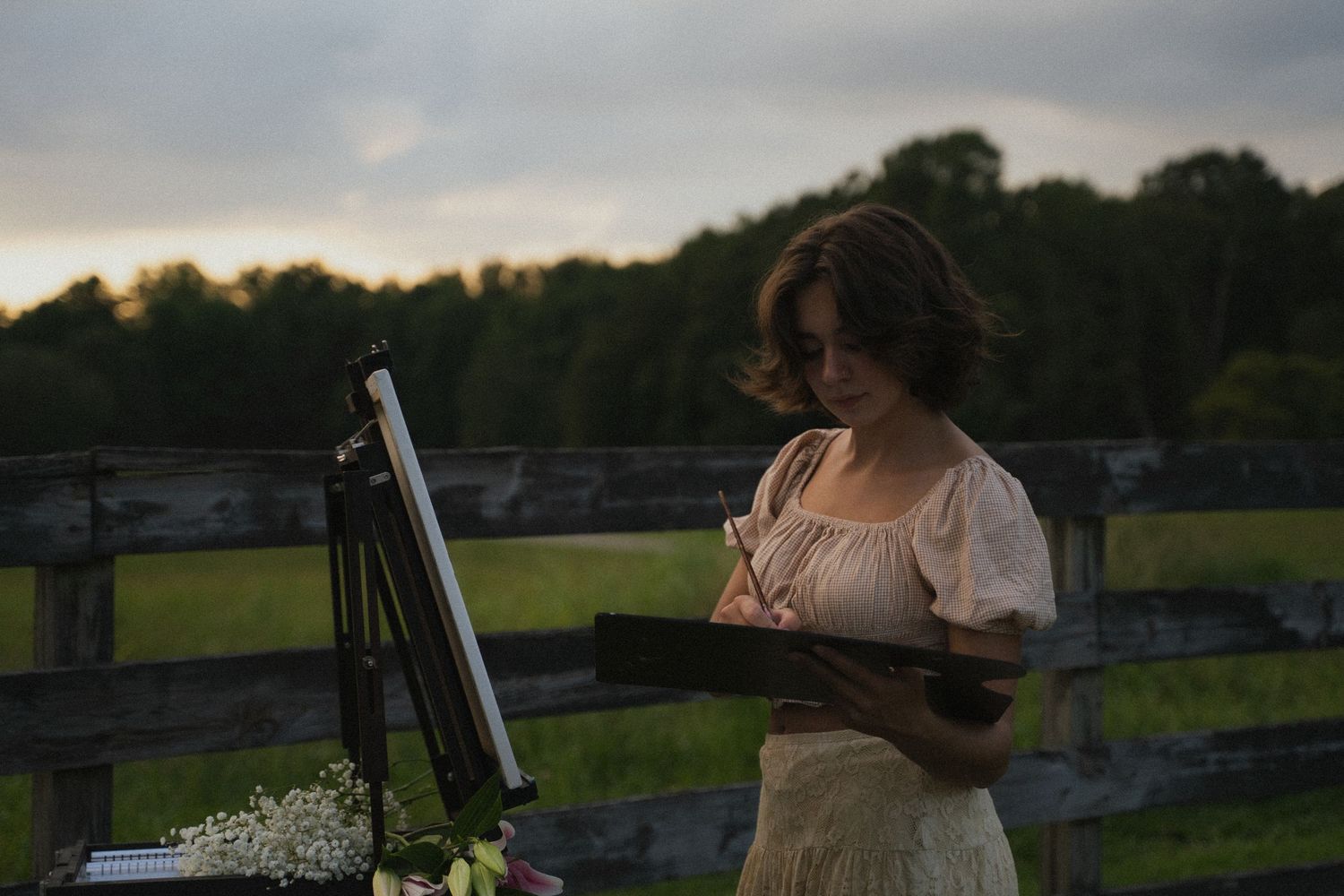 Young woman in vintage dress painting on an easel outdoors near a wooden fence at sunset.