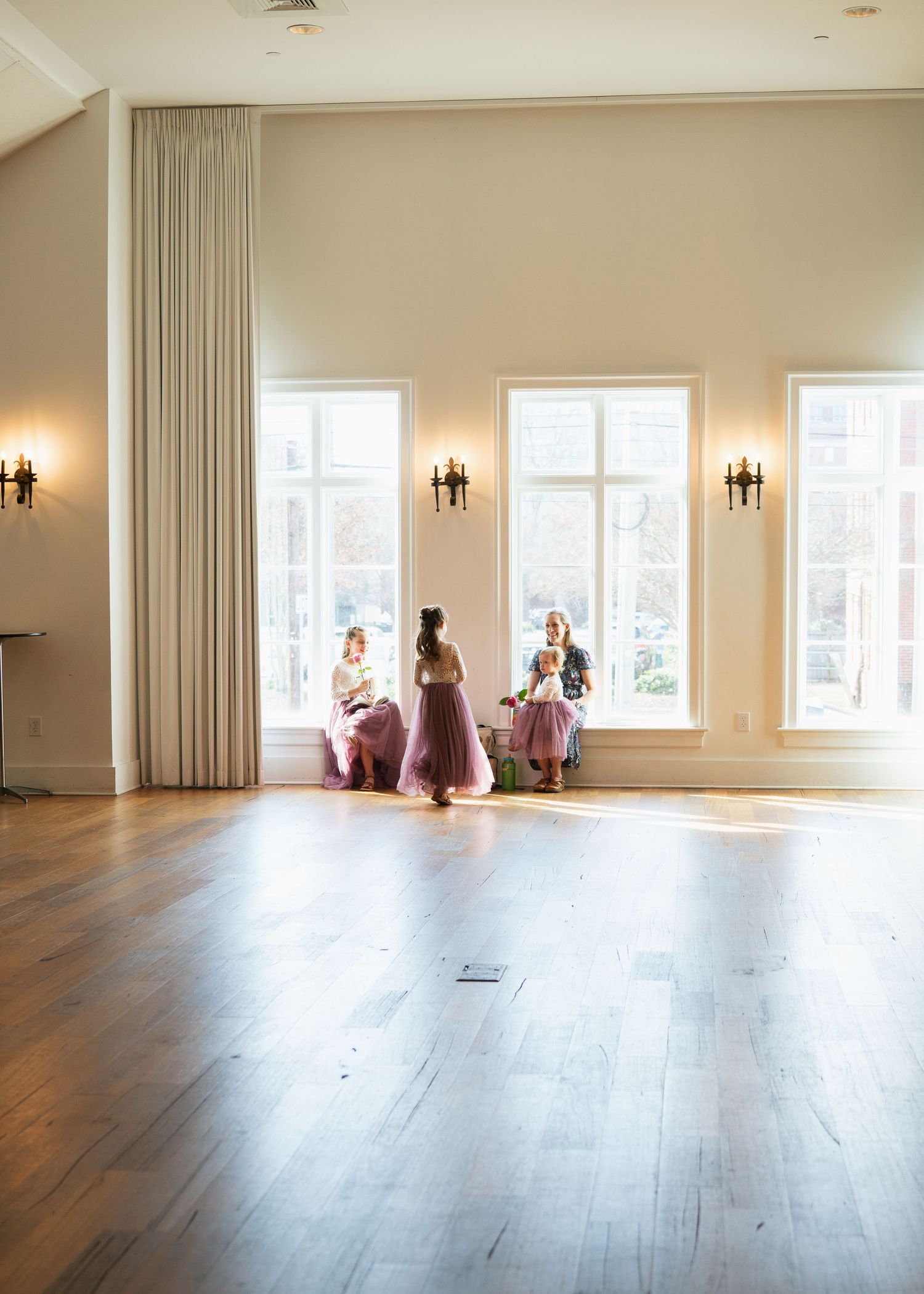 Three children in pink tulle skirts gather by tall windows in bright elegant ballroom.