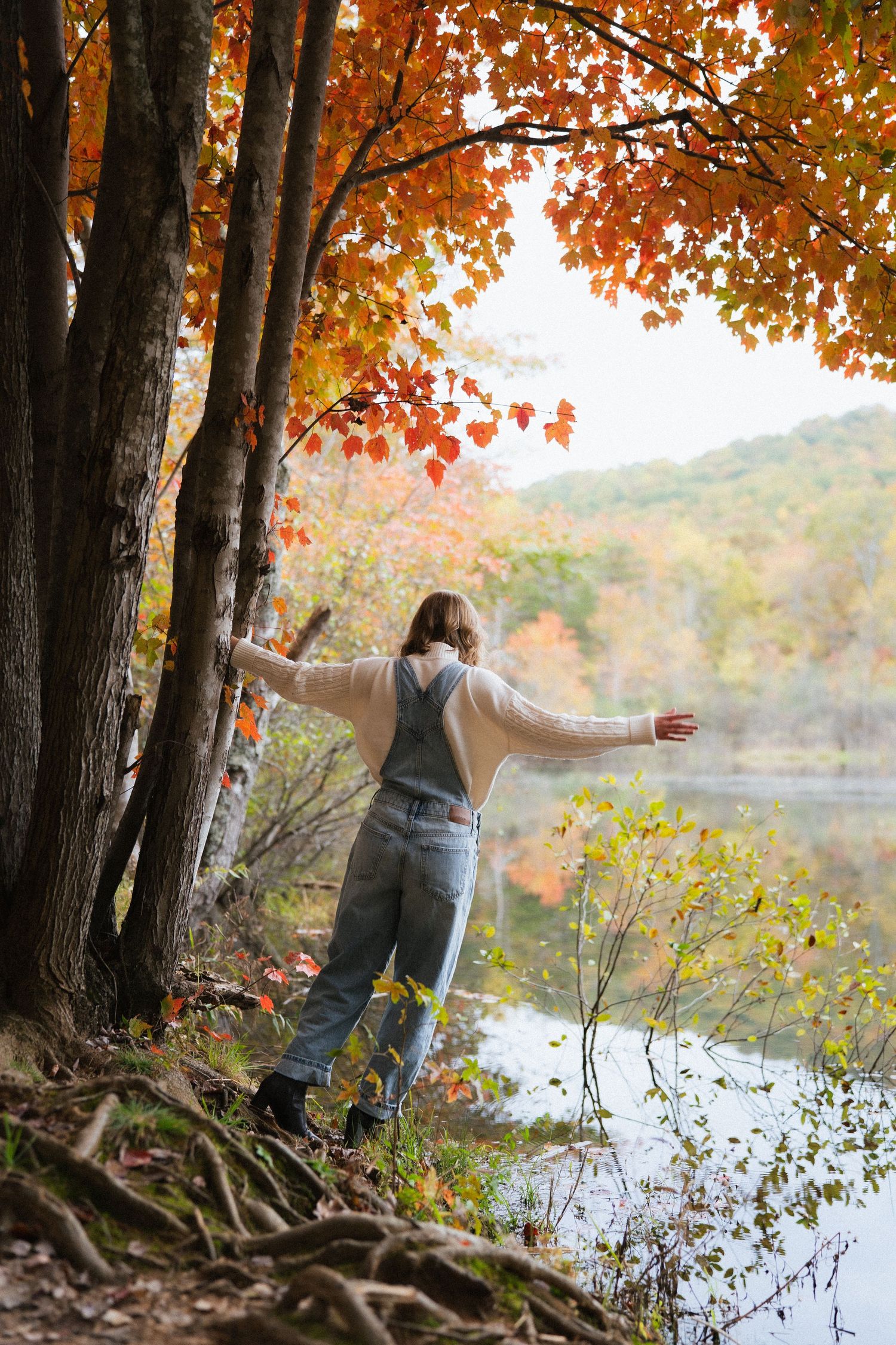Woman with arms outstretched standing by a lake surrounded by vibrant autumn foliage in fall forest.