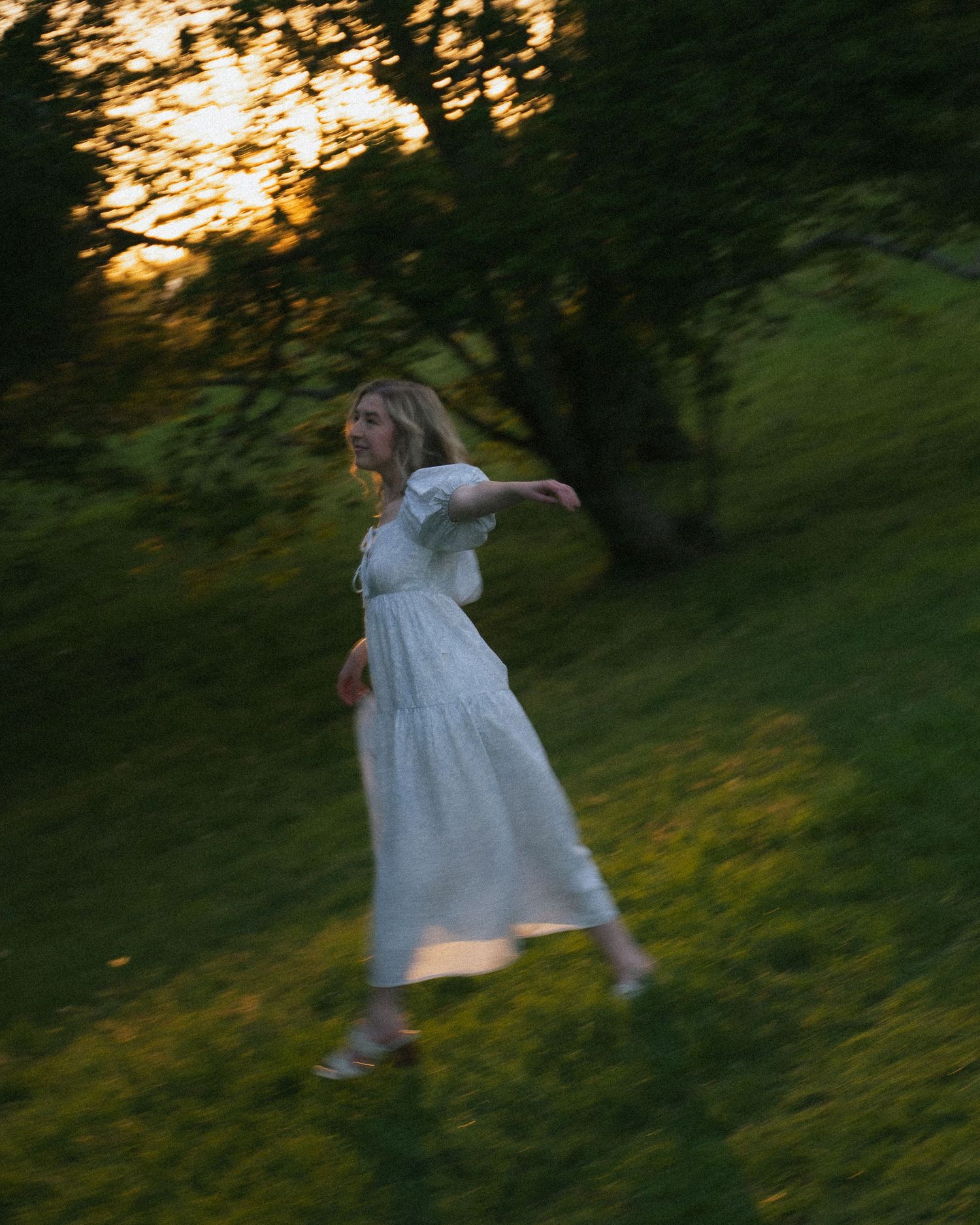 Motion blur of woman in white dress running through green field at sunset.