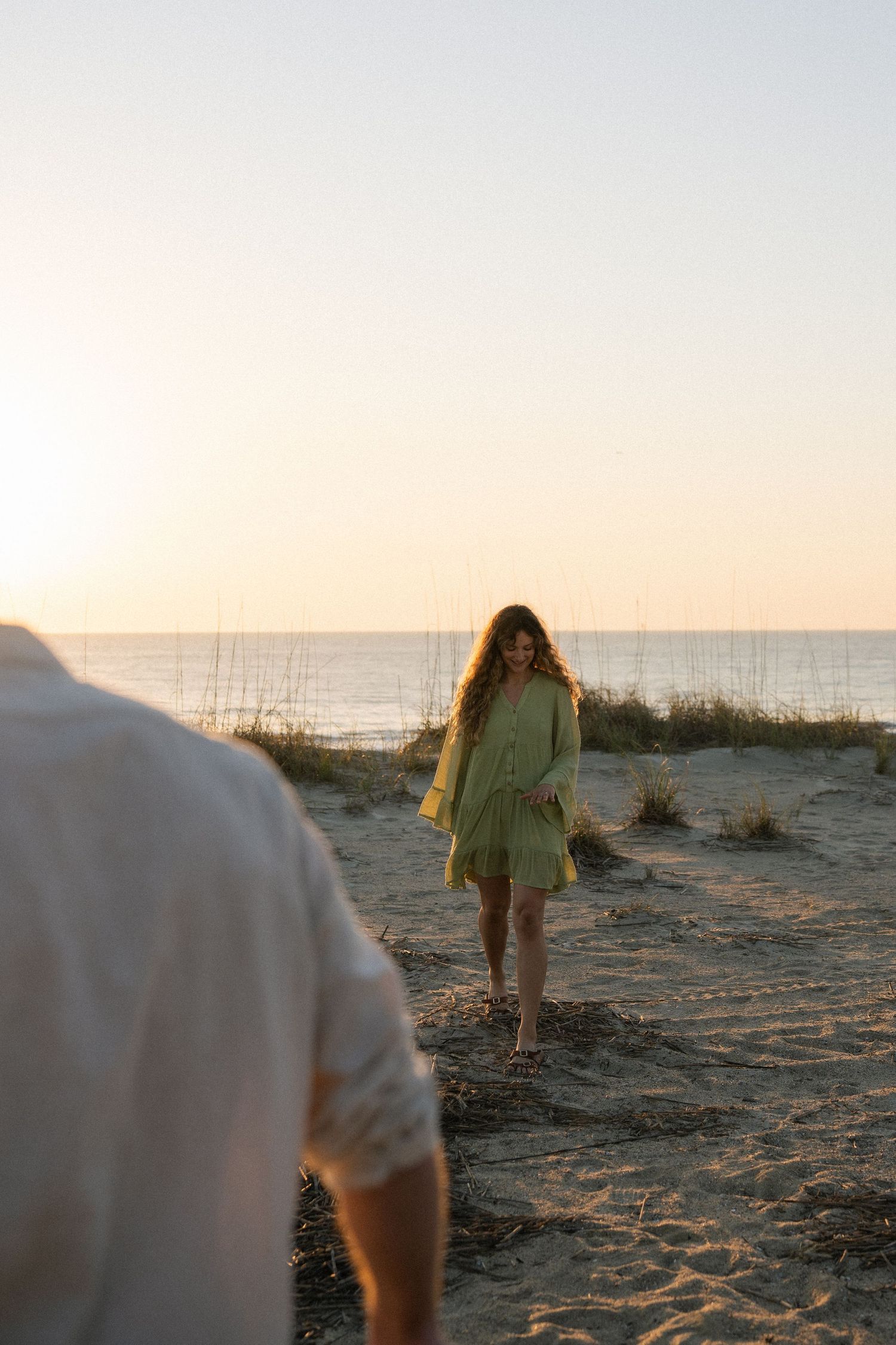Woman in green dress walking toward camera on sandy beach at golden sunset.