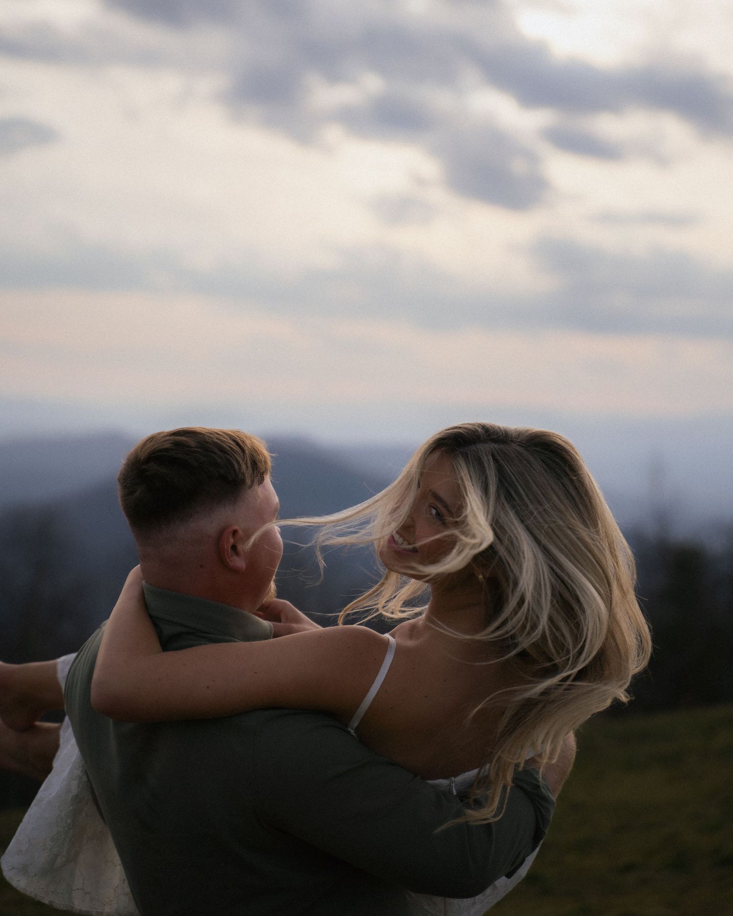 Woman with flowing blonde hair embracing man outdoors under dramatic cloudy sky.