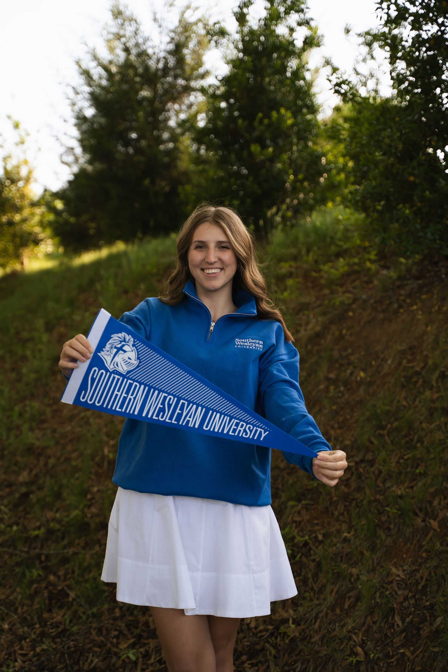 Smiling girl in blue Southern Wesleyan University pullover holding a matching pennant flag outdoors.