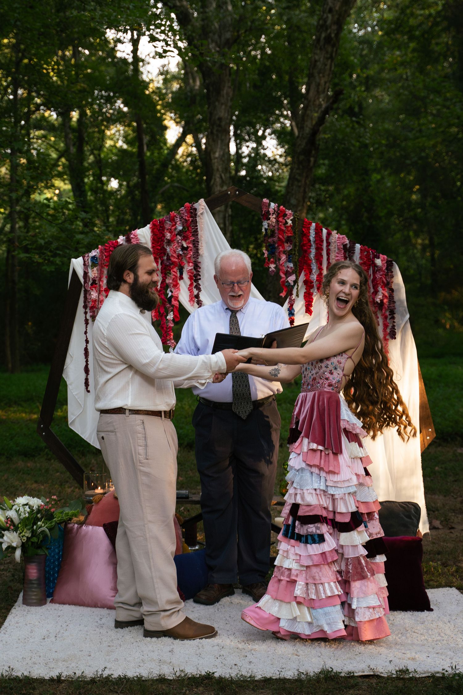 Joyful outdoor wedding ceremony under floral arch with bride in ruffled pink gown and groom in cream suit.