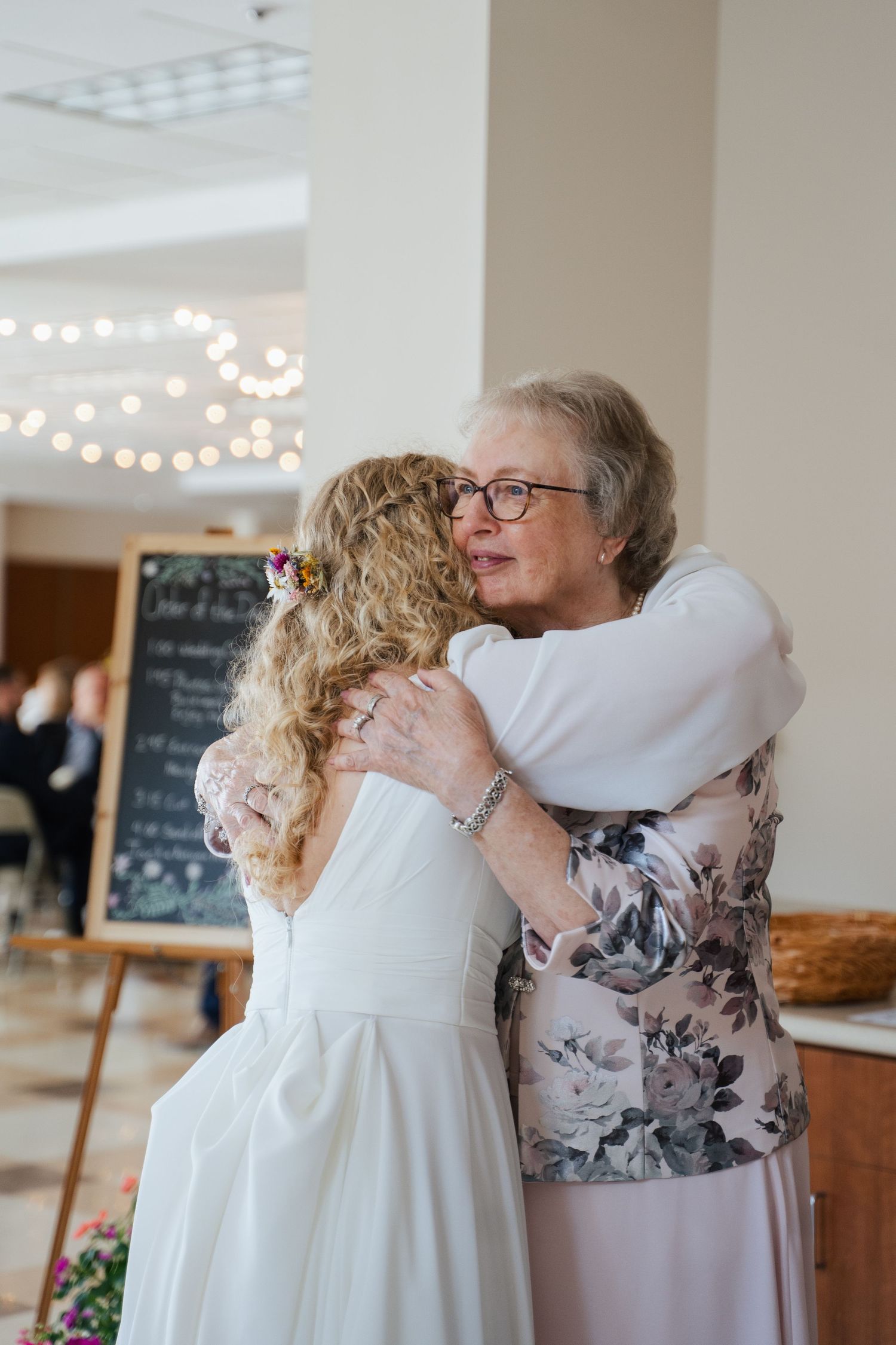 Emotional bride in white wedding dress shares heartfelt hug with older woman in floral jacket at wedding reception.