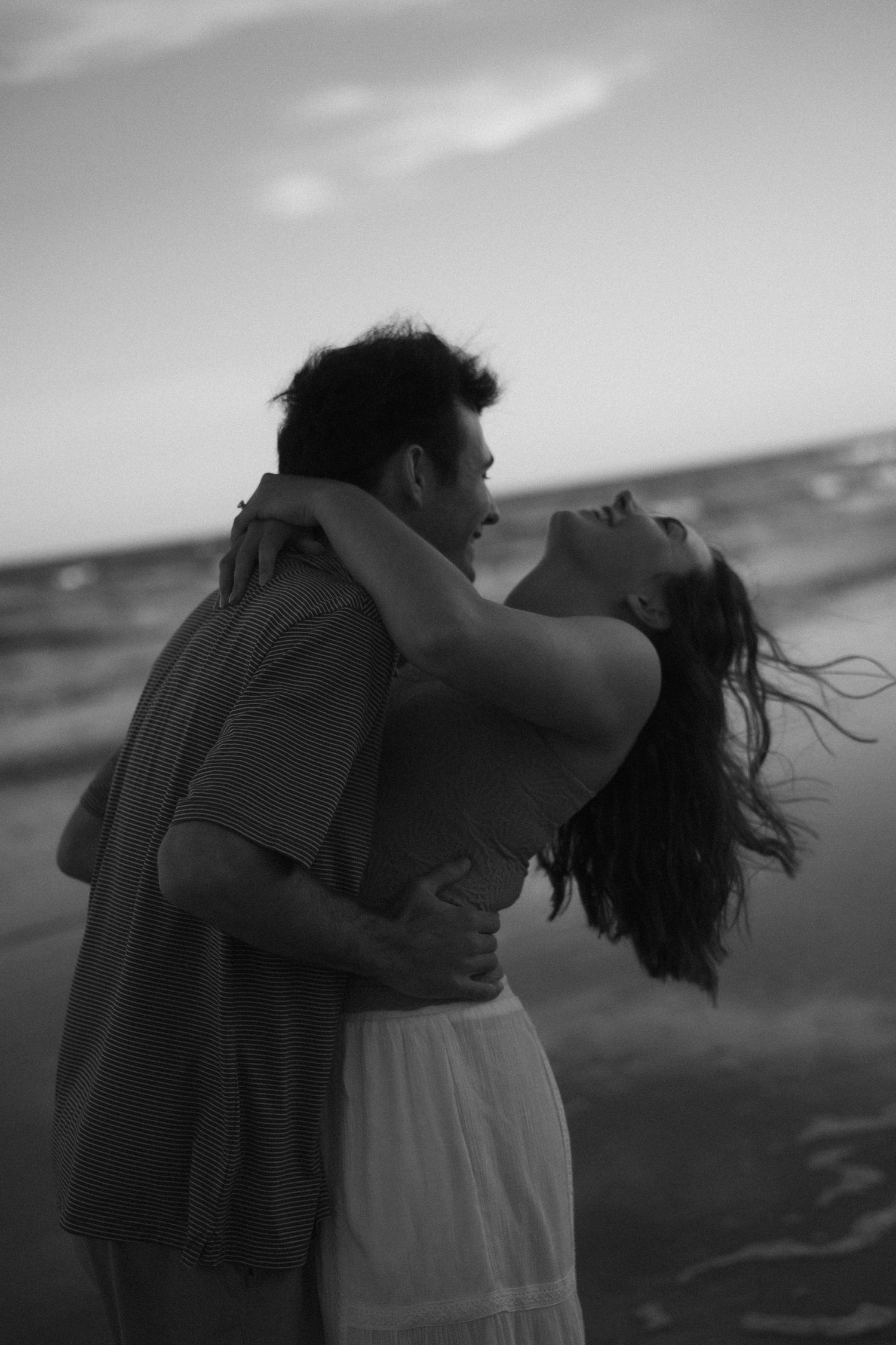black and white photo of a couple embracing on a beach while the woman tilts her head back and the wind blows her hair