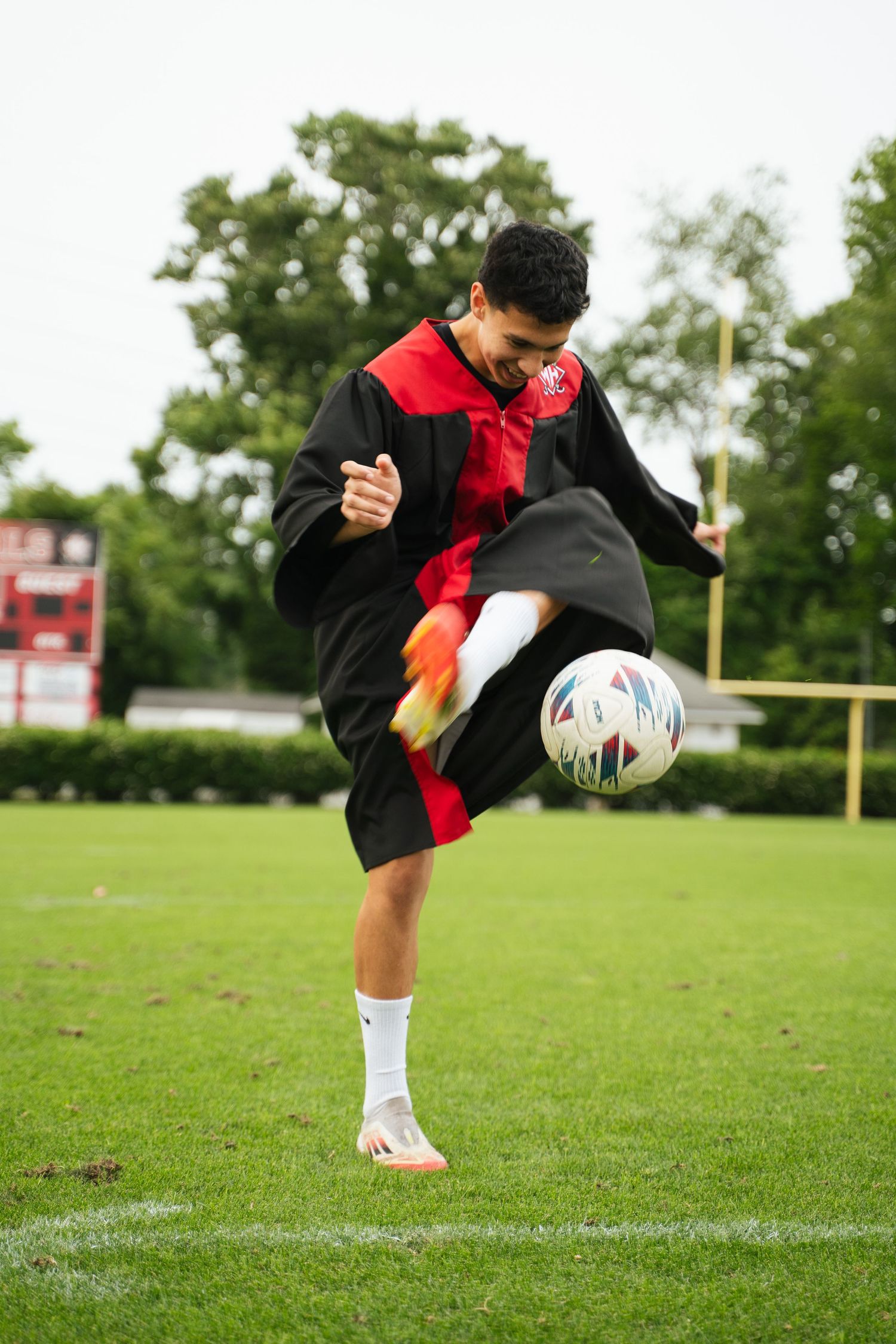 Male graduate in black and red gown kicking a soccer ball on a green sports field outdoors.