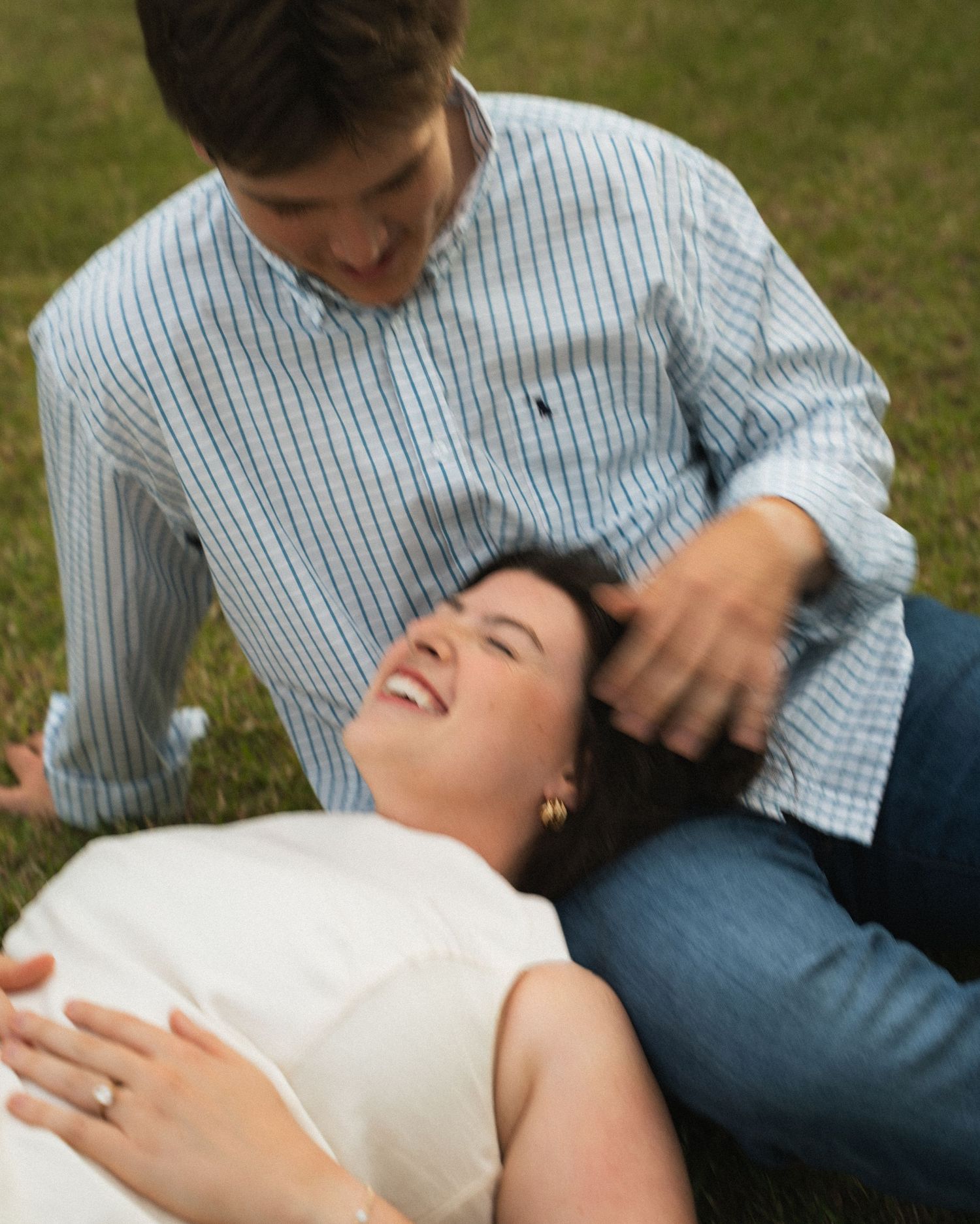 Couple laughing together on grass, woman lying down smiling up at man in light blue striped shirt.